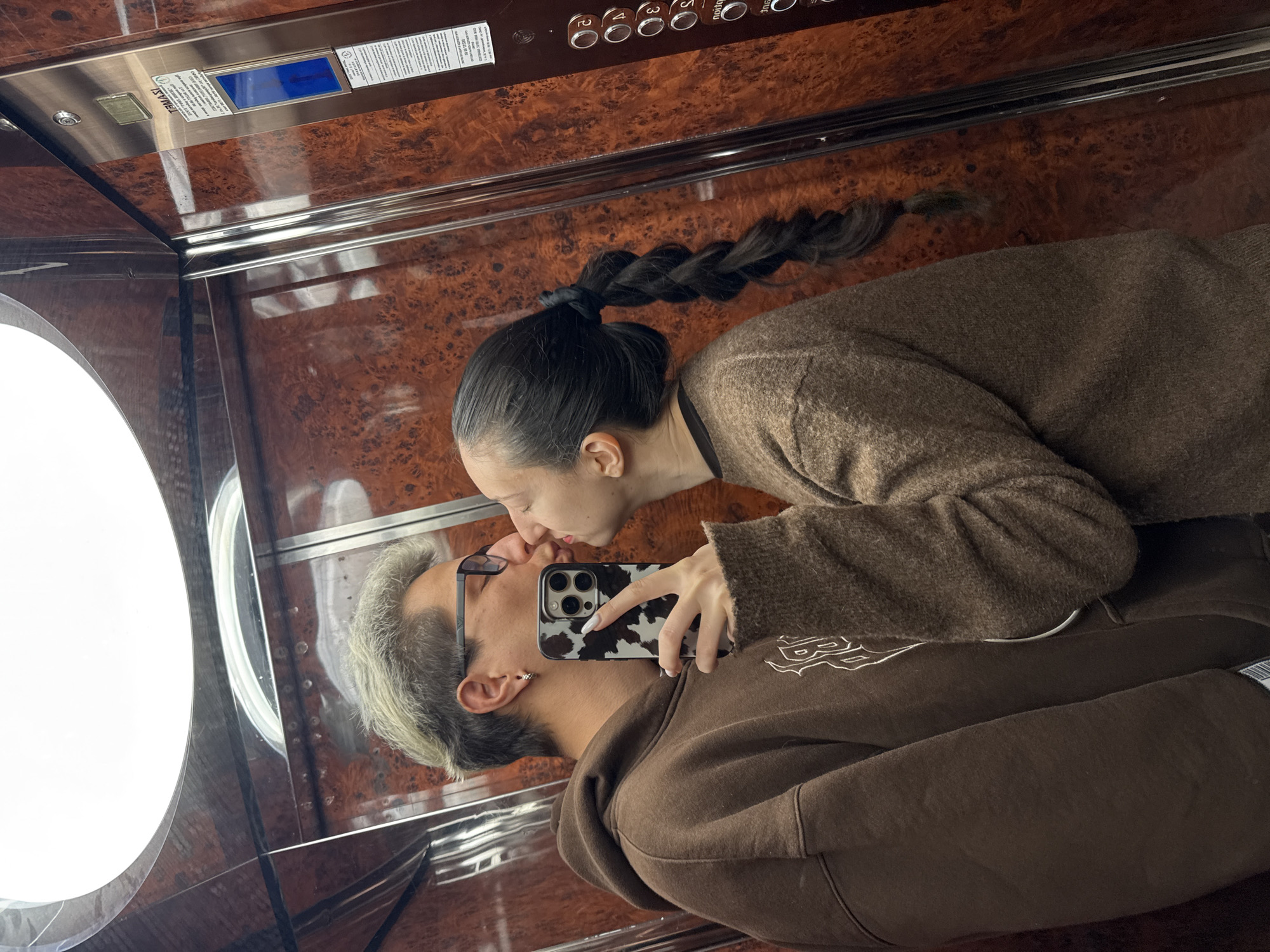 In an elevator with wood-paneled walls and ceiling, a couple is reflected in a mirror taking a selfie while appearing to kiss. The woman has long dark hair in a braid and is wearing a brown sweater. The man has short, light-colored hair, glasses, and is wearing a brown hoodie. The woman is holding a smartphone with a cow-print case. The elevator has a round window and a control panel with buttons. There is no explicit indication of the location being Veliko Tarnovo, Bulgaria.