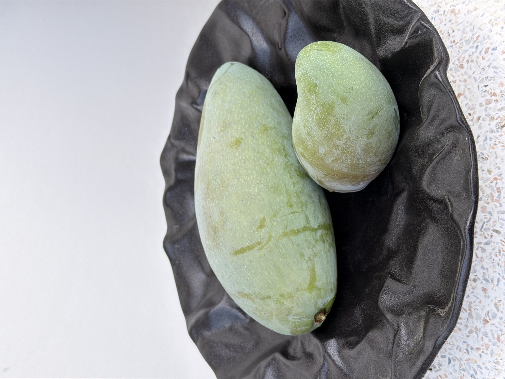Two green mangoes are displayed on a black, wavy ceramic plate, which is situated on a speckled countertop. The larger mango is positioned on the left side of the plate, elongated and slightly curved, with visible water droplets and faint scratches on its skin. To its right, a smaller mango rests, also green with some reddish-brown discoloration near its stem. The background is a plain white wall, providing a stark contrast to the darker elements in the foreground. The image was taken in the Ko Samui District of Thailand.