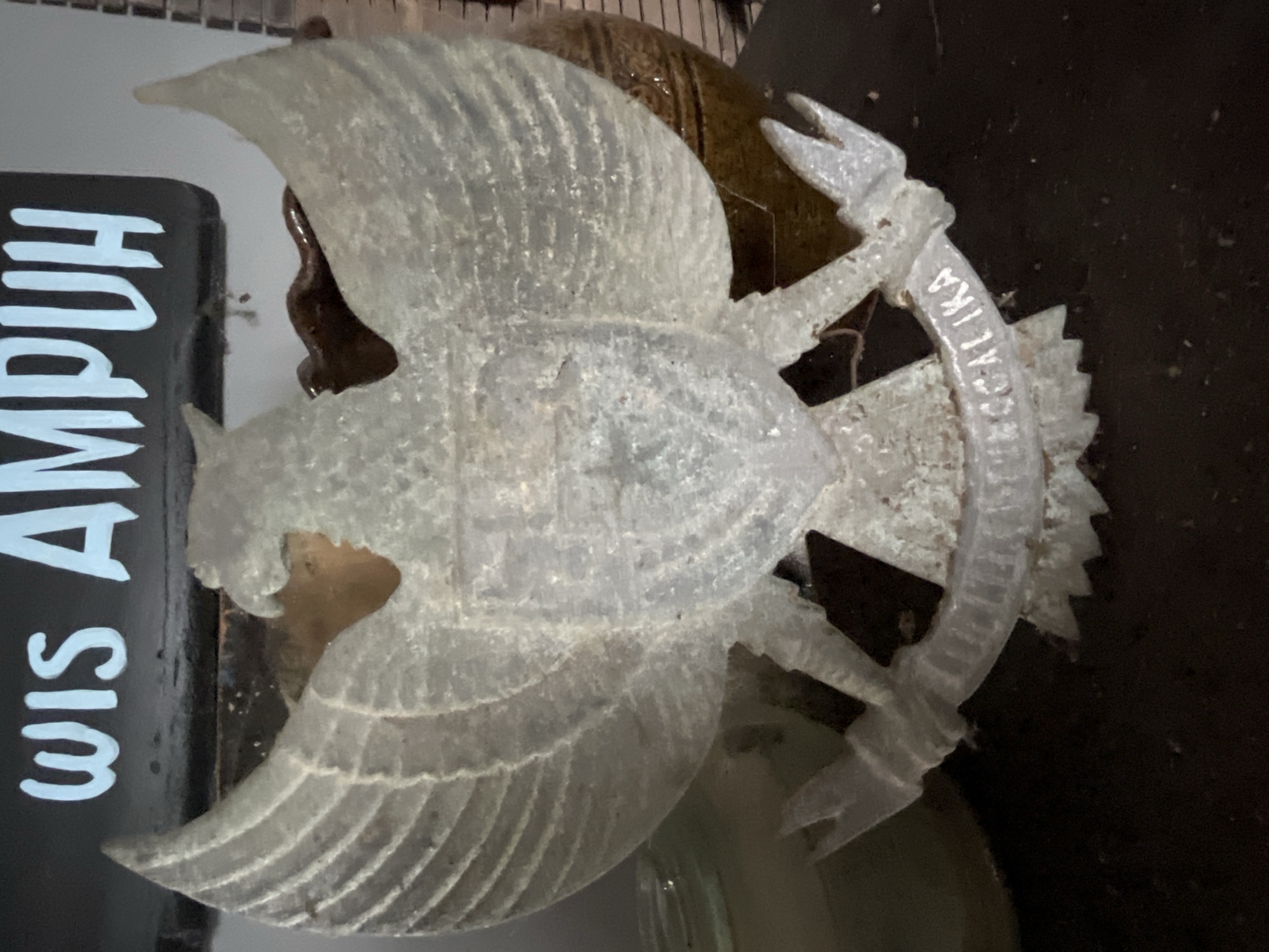 In Tambakreja, Indonesia, a semi-transparent, pale grey emblem of a Garuda, the mythical bird representing Indonesia, is displayed against a dark background. The Garuda is depicted with outstretched wings, a prominent chest, and talons gripping a scroll. The scroll is inscribed with the national motto "Bhinneka Tunggal Ika," meaning "Unity in Diversity," though only the latter part, "GGALIKA," is clearly legible due to the emblem's condition. Above the emblem, white lettering on a dark surface spells out "WIS AMPUH." The emblem appears to be made of a translucent material, possibly glass or a resin, and shows signs of wear, with dust and imperfections visible. The lighting is dim, casting a soft glow on the emblem and highlighting its textured surface. In the background, blurred objects, including what appears to be a ceramic pot and a glass jar, are partially visible. The overall scene suggests a display of national symbolism in a domestic or semi-public setting within Tambakreja.