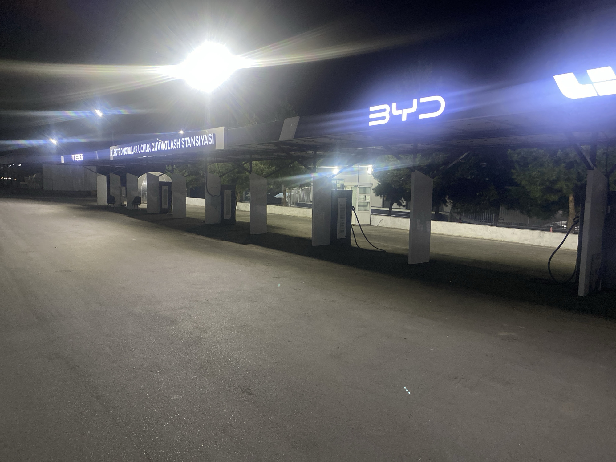 This media file captures an outdoor electric vehicle charging station at night, likely in Temirhoja, Uzbekistan. The scene is illuminated by bright, harsh overhead lights, creating strong glare and lens flare. The primary subjects are the charging stations themselves, characterized by tall, white rectangular structures housing the charging equipment. Cables are visible, suggesting the stations are ready for use.
Above the charging stations, a prominent illuminated sign displays "BYD" in large, white, futuristic lettering. To the left of the BYD logo, another illuminated sign reads "ELEKTROMOBILLAR UCHUN QUVVATLASh STANSIYASI," which translates from Uzbek to "Electric Vehicle Charging Station." This text is also in white, illuminated letters.
The ground is paved with asphalt, appearing wet in some areas, suggesting recent rain or dew. The background features trees and some fencing, indicating an urban or semi-urban environment. The overall atmosphere is functional and modern, with no people visible, suggesting the station is either closed for the night or automated. The time of day is clearly night due to the darkness and artificial lighting. The weather appears to be clear, with no indication of rain or strong wind. The lighting creates a somewhat stark and high-contrast image.