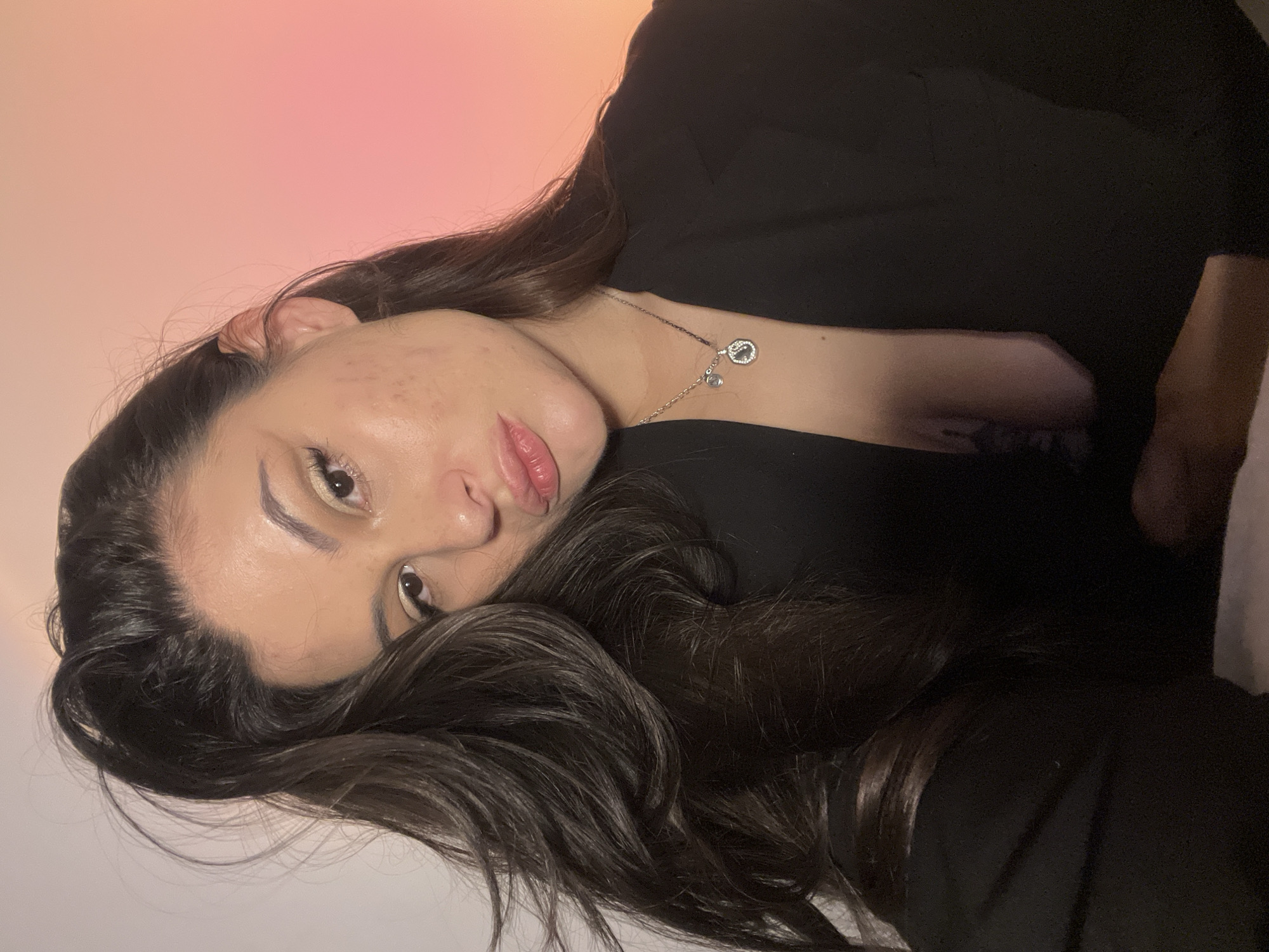 The image depicts an adult female in an indoor setting located in the city of Kiryat Bialik, Israel. The subject has long, dark hair, styled with volume and some strands appearing swept. Her face is directed towards the camera, exhibiting a direct gaze and slightly pursed lips. Visible makeup includes light-toned eyeshadow and lip color. Skin texture, including minor blemishes, is evident on her cheeks. She wears a black garment, resembling a blazer or top, with an open neckline that exposes a portion of her upper chest. A silver-colored necklace with two pendants, one small and circular, the other larger with an open, embellished design, rests on her chest. A dark tattoo is partially visible on her upper chest. The background consists of a plain wall, white on the left side, transitioning to a soft orange-pink gradient on the right side, suggesting artificial mood lighting.