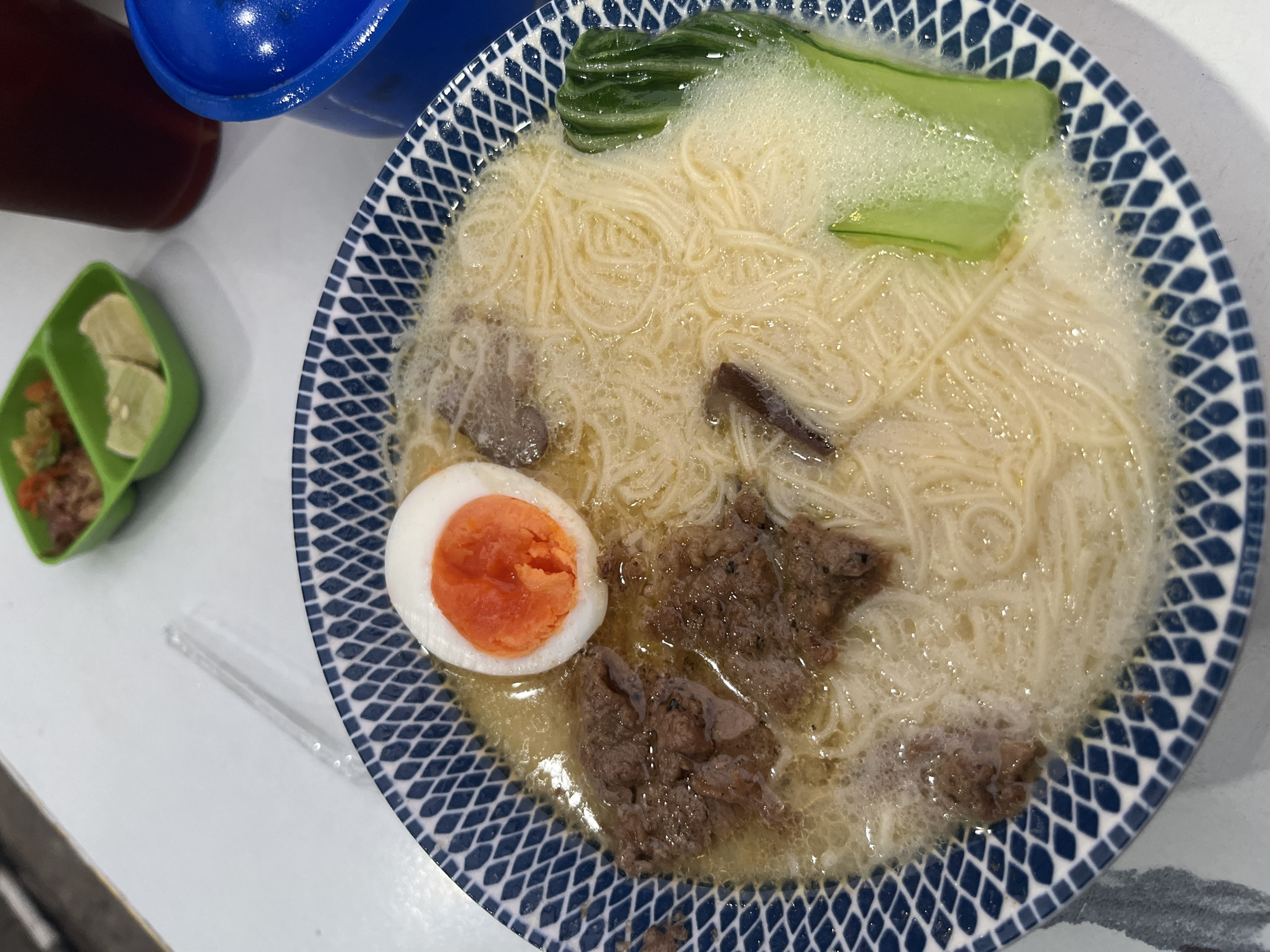 A bowl of ramen with noodles, a halved soft-boiled egg, and meat is presented on a white table. A small green container with lime wedges and a condiment sits in the upper background. A dark beverage and a blue lid are also visible. The ramen bowl has a blue and white patterned rim. The scene appears to be in a restaurant.