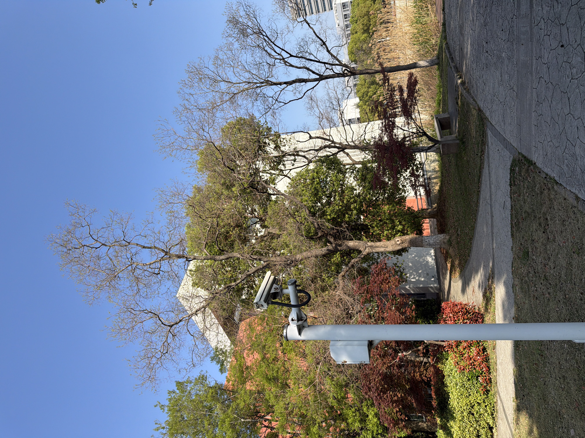 The image depicts an outdoor scene in Jiangning District, China, characterized by a clear blue sky above. Dominating the foreground is a white pole supporting two surveillance cameras. The background features a mix of trees, some with bare branches, while others display green and red foliage. The architecture includes a white building partially obscured by the trees. A paved walkway runs along the right side of the image, with patches of grass visible between the walkway and the vegetation. A concrete bench is situated adjacent to the walkway.