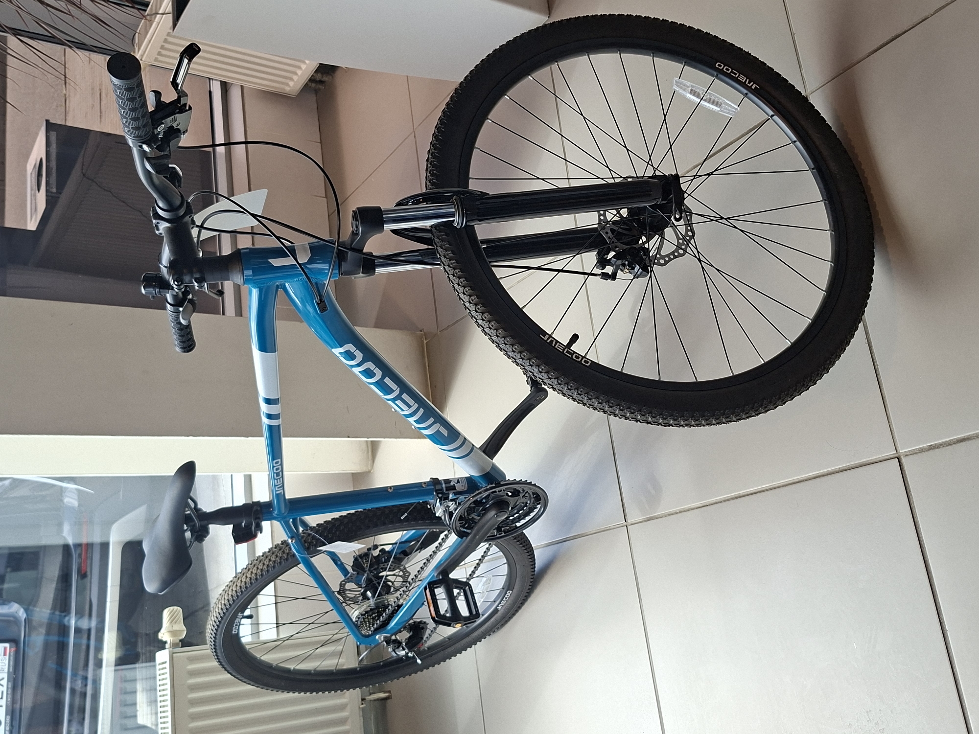 The image shows a blue and white JNECOO brand bicycle, likely a mountain bike, indoors. The bicycle is positioned in a well-lit room with a tiled floor. The background includes a radiator, a window reflecting some objects outside, and white partition walls. The bicycle features black handlebars with grips, brake levers, and a bell, a black saddle, a blue frame with white accents, black wheels with tires and spokes, a crankset with pedals, and a disc brake system on the front wheel. The brand name "JNECOO" is visible on the frame and tires. There are no people visible in the image. The scene is consistent with a retail display or a storage area in a bicycle shop. It is not possible to determine the specific location in the city of Izhevsk, Russia from the visual information provided.