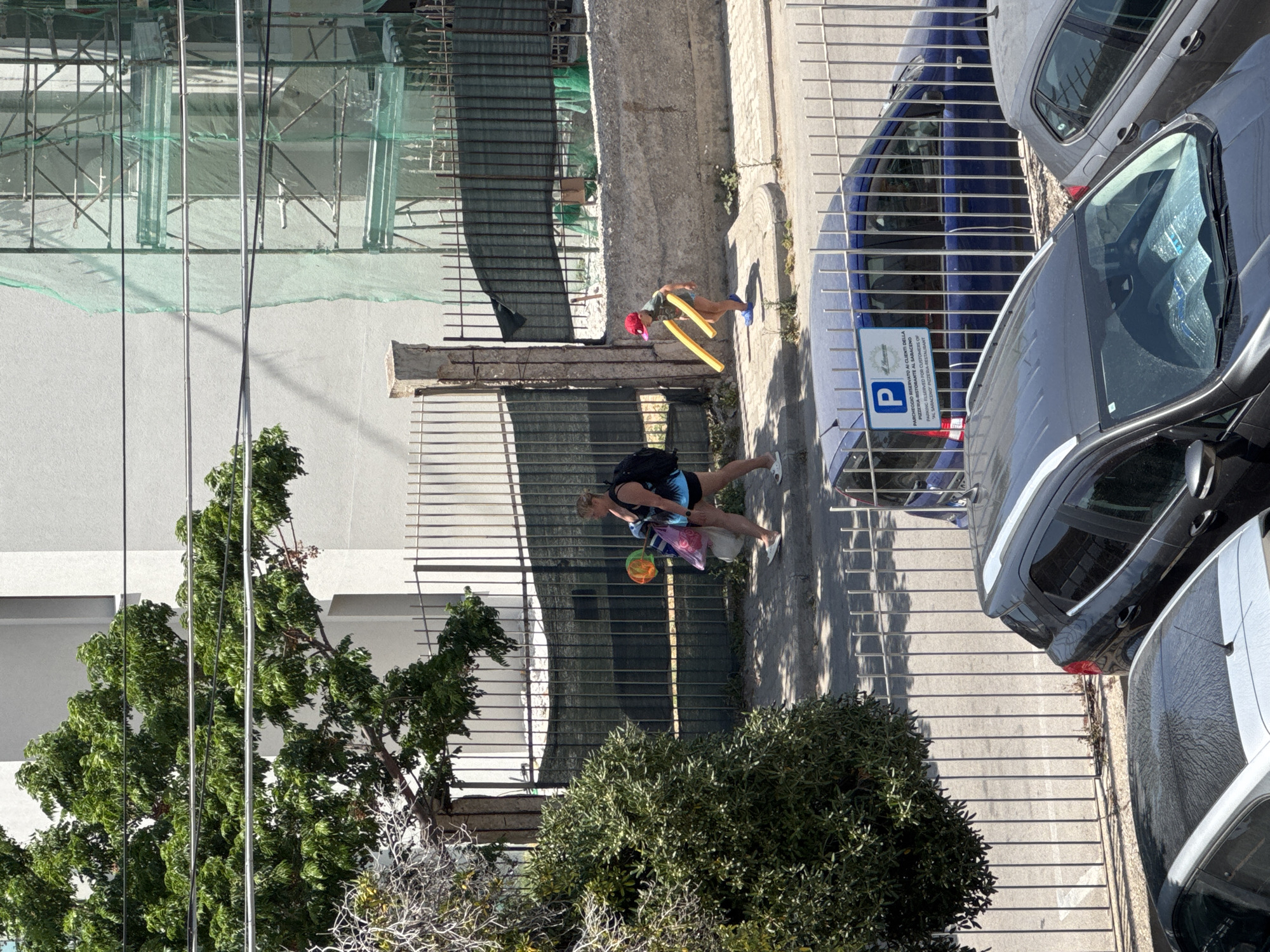 An overhead view captures a sunlit street scene in Capaci, Italy. An adult, likely a woman, with a dark backpack and carrying colorful plastic bags and a small, colorful ball, walks on a concrete sidewalk. Ahead of her, a child, wearing a red hat and blue shoes, carries two long, yellow pool noodles. Both appear to be heading towards a beach or pool, indicated by their accessories.
To their left, a metal fence separates the sidewalk from a construction site, identifiable by scaffolding and green safety netting covering a building under renovation. A large, leafy green tree partially obstructs the view of another building further left. On the right side of the sidewalk, several cars are parked alongside the curb, including a dark gray SUV in the foreground, a blue car, and part of a white car.
A white parking sign is visible next to the parked cars, featuring a blue 'P' symbol. The text on the sign, in both Italian and English, reads: "PARCHEGGIO RISERVATO AI CLIENTI DELLA PIZZERIA-RISTORANTE AL SARACENO" and "PARKING RESERVED FOR CUSTOMERS OF 'AL SARACENO' PIZZERIA-RESTAURANT". Strong, defined shadows cast by the fence, cars, and people indicate bright, sunny weather, likely mid-morning or afternoon. The overall scene depicts a typical summer day in a Mediterranean town.