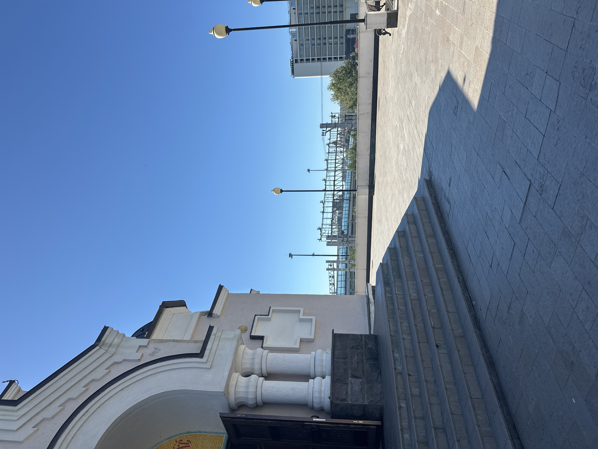 The image captures an outdoor scene in the city of Sirius, Russia. In the foreground, a set of stone steps leads up to the entrance of a pale pink building with white architectural details, including fluted columns and a cross-shaped emblem. The entrance is marked by an open dark wooden door. To the right of the building, a paved courtyard stretches out, marked by a sharp shadow cast by an unseen object.
Beyond the courtyard, a white railing runs along the edge of an elevated area. In the midground, a body of water is visible, crossed by a large industrial structure that appears to be a bridge or a port facility, with intricate metalwork and electrical lines. Several lampposts with decorative yellow tops are spaced along the elevated area.
Further in the background, a modern, multi-story white building stands tall, likely a hotel or residential complex, with many visible windows. The sky is a clear, bright blue. The overall impression is one of a sunny day, with a mix of historical architecture and modern urban infrastructure. There are no visible people in the scene.
