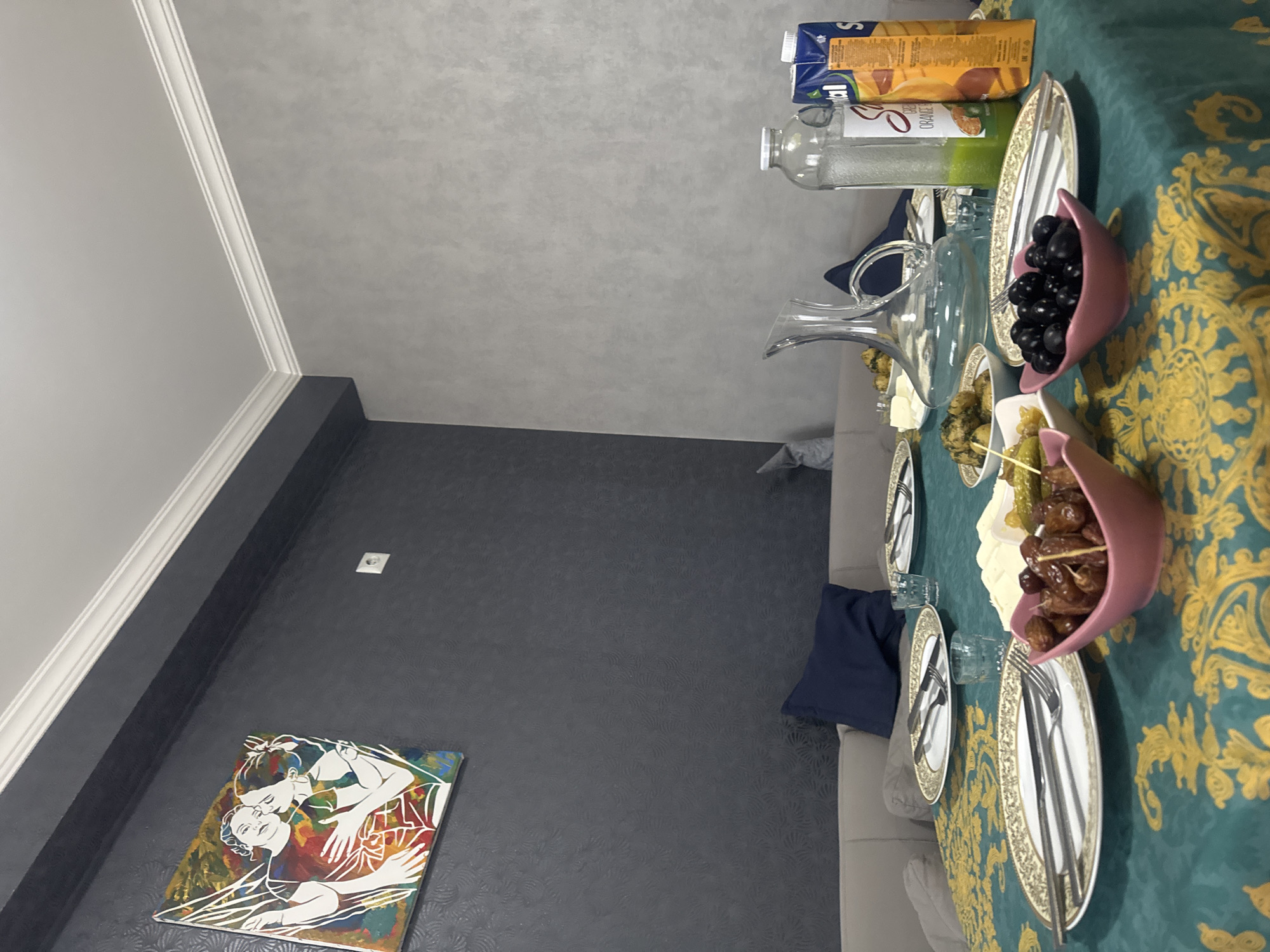 This indoor image captures a dining setting, likely in Tbilisi, Georgia, prepared for a meal. A table in the foreground is draped with a vibrant teal tablecloth featuring an elaborate gold damask pattern. It is set with several white plates with decorative gold rims, cutlery (forks and knives), and clear drinking glasses.
A variety of appetizers and beverages are arranged on the table. A clear glass decanter is prominent, alongside two juice cartons, one visibly labeled "ORANGE". Two distinctive pink, boat-shaped bowls are filled with black olives and dates. Other small white bowls contain assorted items, including what appears to be cubed cheese or butter, pickled gherkins, and a green, possibly herb-based or seasoned vegetable dish.
The background shows two distinct wall finishes: a light grey wall with a subtle texture meeting the ceiling with white decorative molding, and a darker grey wall with a faint, repeating circular or fan-like pattern. A framed piece of art hangs on the darker wall, depicting stylized white outlines of two intertwined figures, possibly embracing, against a vivid, abstract background of red, blue, yellow, and green. A white electrical outlet is also visible on this wall. A light-colored sofa or couch with a dark blue throw pillow is partially visible to the right. The scene suggests a cozy and inviting atmosphere, possibly for an evening meal or gathering. No people are visible, and the time of day is inferred to be indoors with artificial lighting.