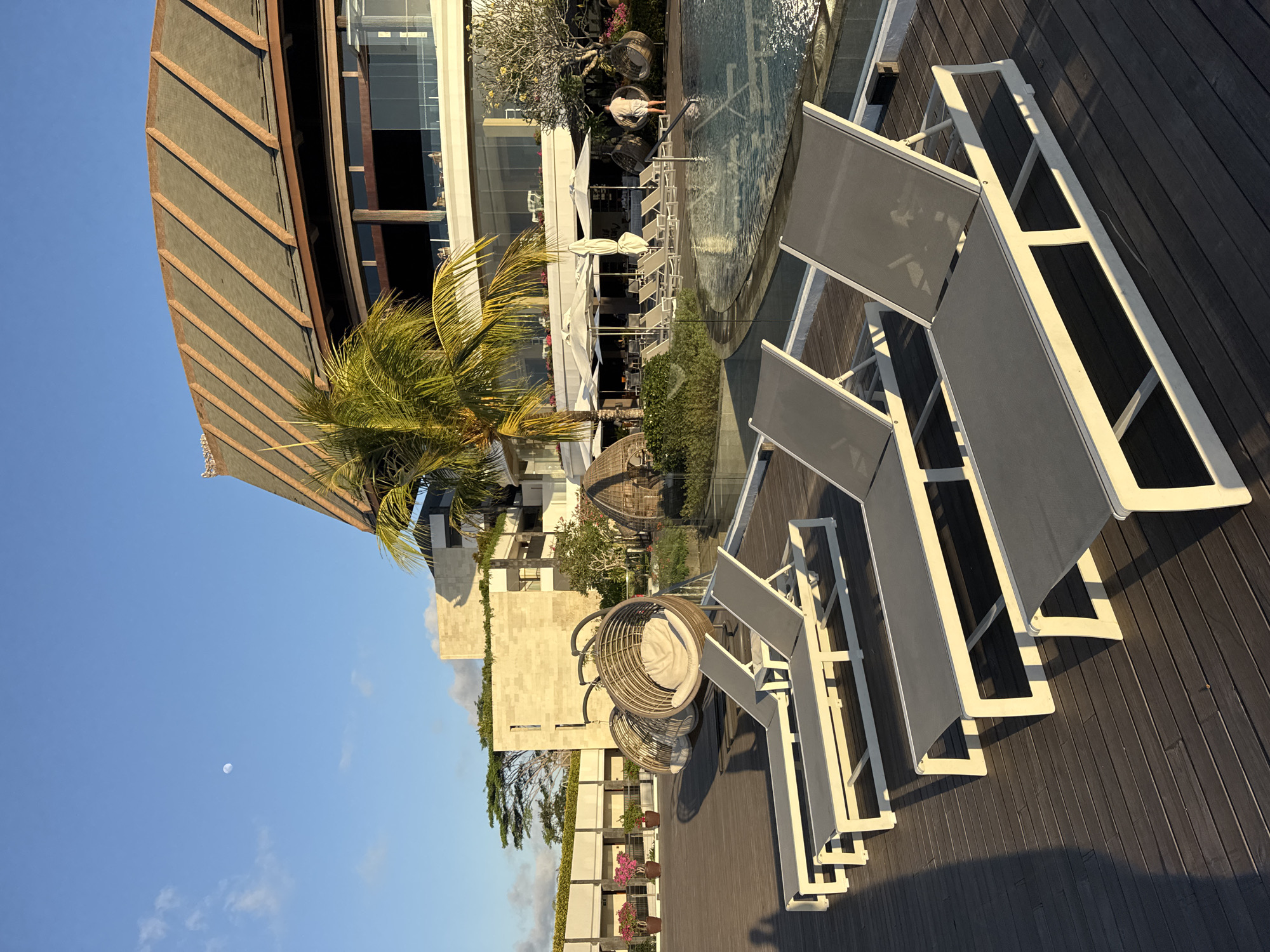The image depicts a resort scene in Kuta Selatan, Indonesia, likely at sunset or sunrise given the warm lighting and long shadows. In the foreground, a wooden deck is occupied by several empty, grey-upholstered lounge chairs. To the left of the chairs, the shadow of a person is cast on the deck. Behind the lounge chairs, a pool area is visible, featuring lounge chairs, umbrellas, and what appears to be a swimming pool. Further back, a large building with a distinctive sloping roof, possibly a resort or hotel, stands against a blue sky with a visible moon. The building is surrounded by greenery, including palm trees and flowering plants. Two hanging rattan chairs are also situated on the deck. A single person, wearing light-colored shorts and a shirt, is standing near the pool.