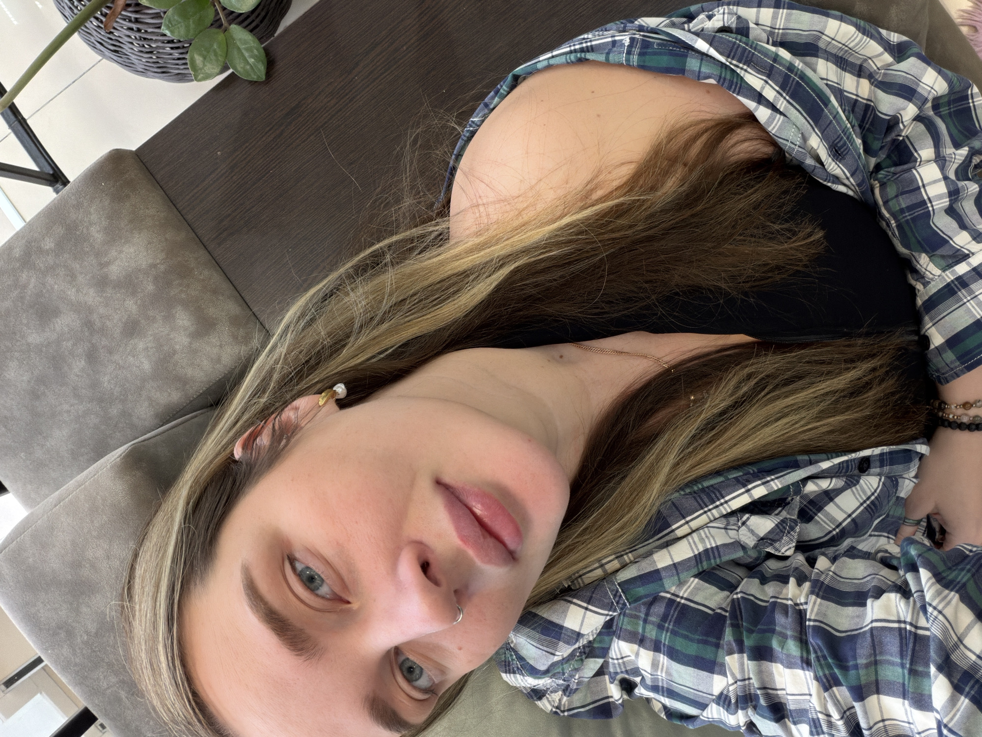 A medium shot shows a woman lying on a gray sofa or chair. She wears a blue and green plaid shirt over a black top. She has long blonde and brown hair. She has a nose ring and an earring. The background includes a dark brown table and a potted plant. The location is Chaikovsky, Russia.