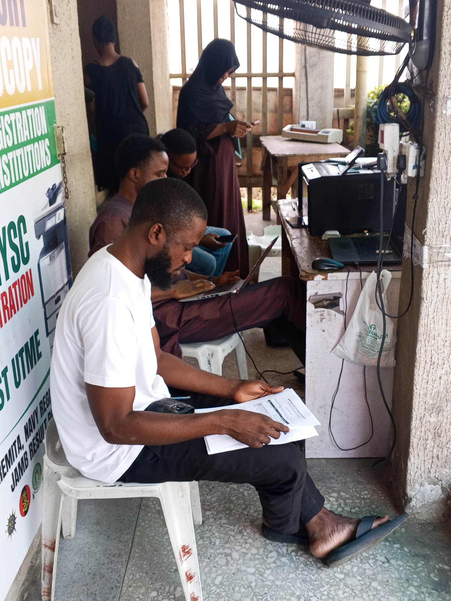 In Lokoja, Nigeria, a group of individuals are gathered in an outdoor area, likely a commercial space or roadside stall, characterized by concrete flooring, a textured wall, and a slatted metal barrier. The scene appears to be related to an administrative or registration service.
A man with a beard, wearing a white t-shirt and black pants, is seated on a white plastic chair. He is holding and examining documents, suggesting he is either waiting for a service or performing administrative tasks. His posture and focus indicate engagement with the papers.
To his left, a sign with text in yellow and green is visible, featuring the words "COPY," "REGISTRATION," and "INSTITUTIONS," implying the services offered at this location.
In the background, several other individuals are present. A woman in a dark headscarf and a long brown garment is standing and appears to be interacting with a mobile phone or device. Another man is seated behind her, also looking towards a laptop. Further back, another person is partially visible, also seated.
The environment includes various objects associated with technology and service provision. A laptop is open on a desk, accompanied by a computer mouse. A printer and a laminating machine are also present on the desk, suggesting document processing services. Electrical outlets and cables are visible, indicating the use of electronic equipment. A large ceiling fan is mounted overhead, providing ventilation. A white plastic bag with text and logos is hanging from a utility pole. The overall atmosphere suggests a functional, albeit somewhat utilitarian, service point.