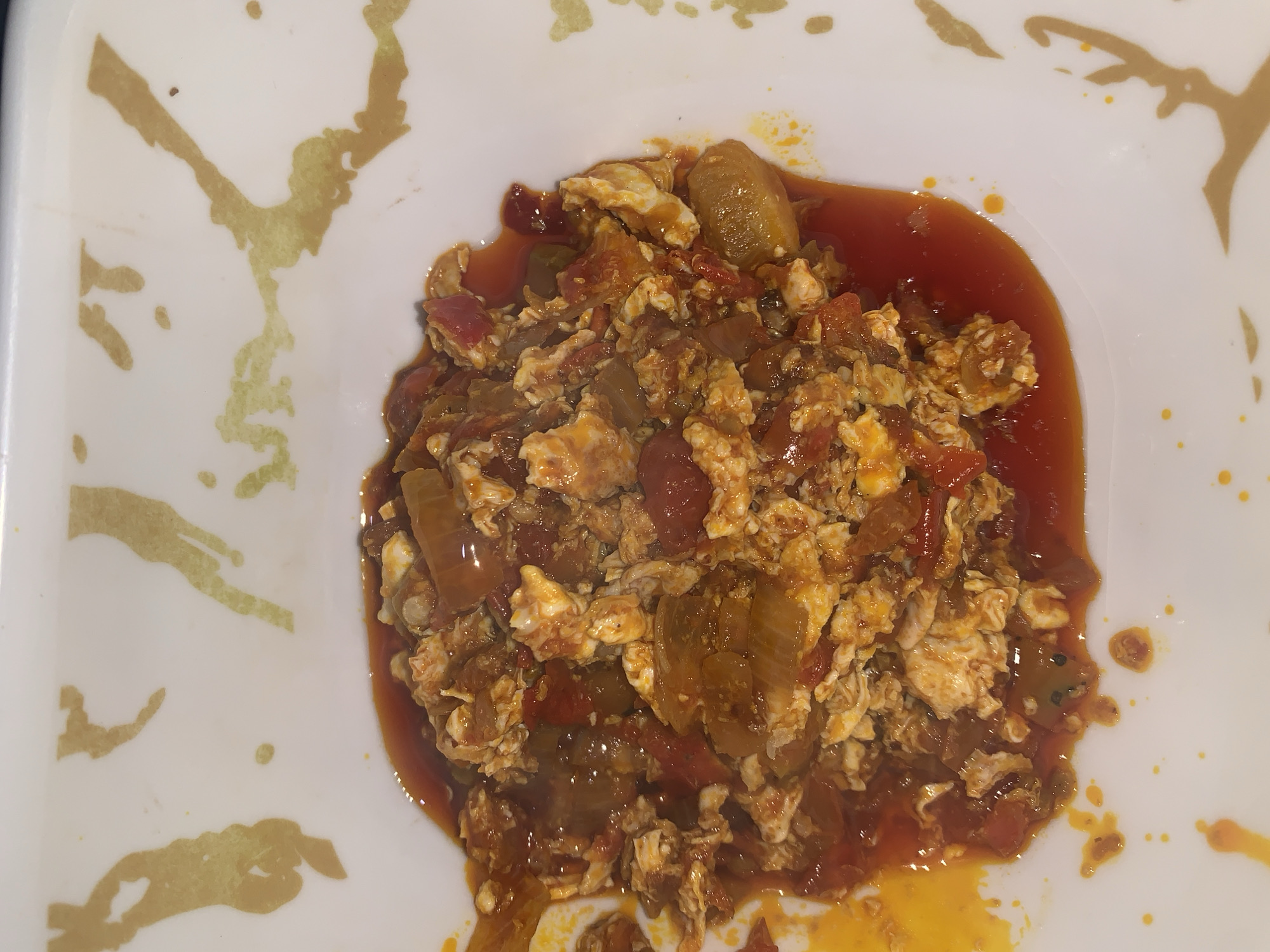 A close-up, overhead shot shows a white bowl with a gold abstract design on its rim filled with a dish of scrambled eggs with tomatoes and onions in a reddish-orange sauce. The food is in the center of the bowl, with some sauce spilling over the sides. The background is out of focus and appears to be a light-colored surface. The image quality is slightly grainy and the lighting is dim. There are no people or other objects visible in the scene. The location is specified as Rafin Par, Nigeria.