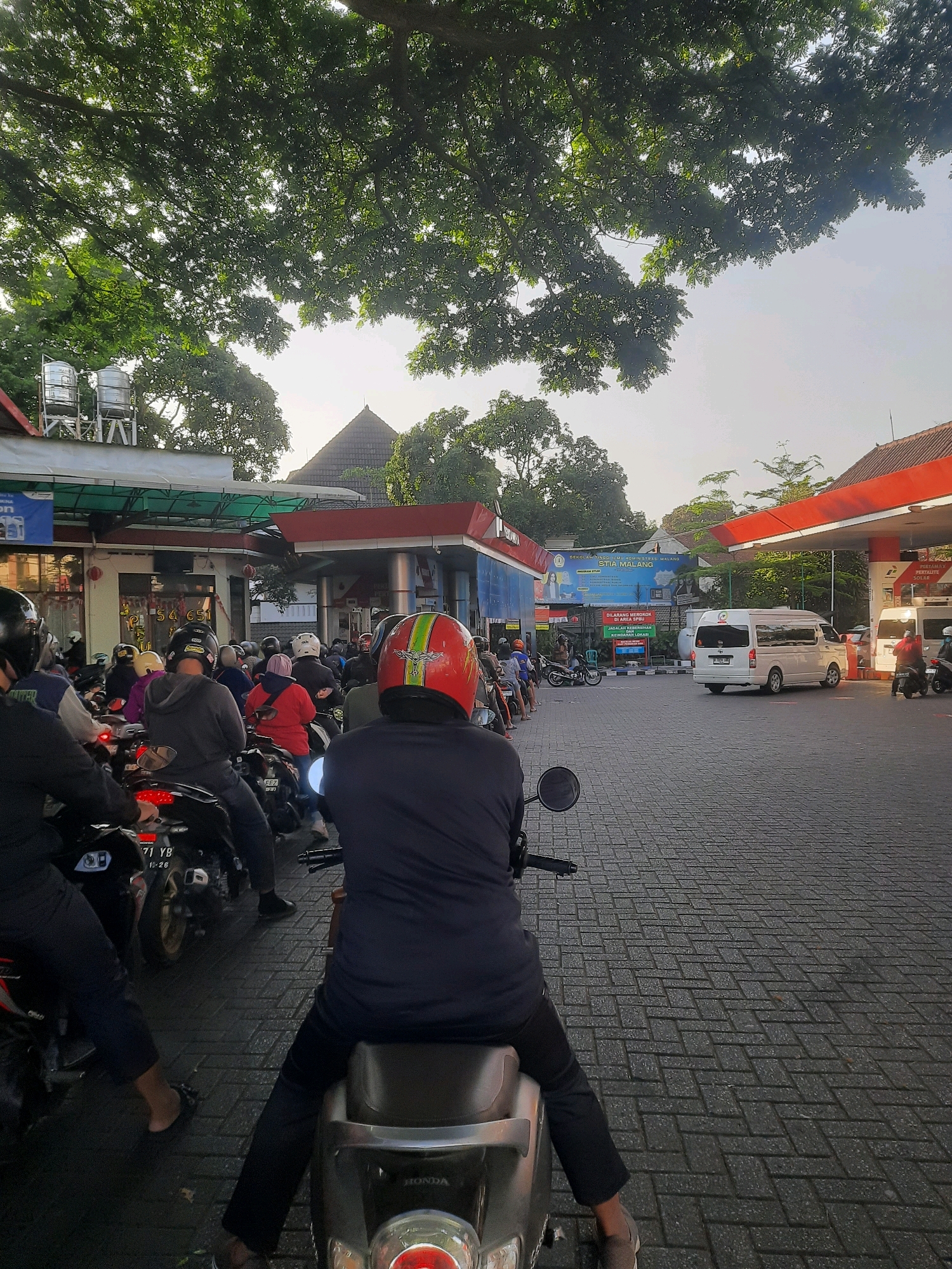 The image depicts a busy gas station in Malang, Indonesia, with a long line of motorcycles and a few cars waiting to refuel. The time of day appears to be late afternoon or early evening, judging by the soft, warm light filtering through the trees. The gas station has red and white awnings, typical of many in Indonesia.
In the foreground, a person on a scooter, facing away from the camera, is part of the queue. They are wearing a black jacket and pants, and a red and green helmet. Behind them, more scooters and riders fill the frame, creating a dense line stretching towards the pumps.
Further into the scene, a white van is visible, also waiting. The gas station buildings are visible in the background, along with trees and a hint of residential buildings or other structures. The ground is paved with interlocking bricks, common in Indonesian cities. The overall impression is one of a typical, bustling day at a public fueling station.
