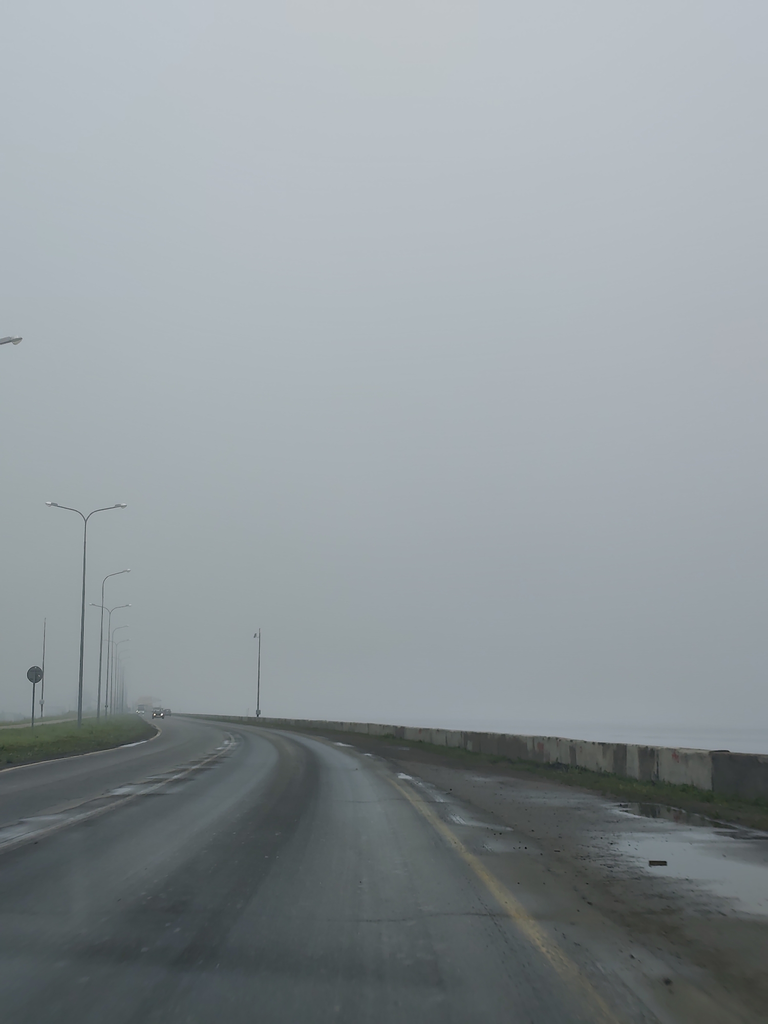 The image captures a foggy, wet, and overcast scene along a curved roadside, likely viewed from a moving vehicle. The dominant feature is a long stretch of dark, wet asphalt road, showing reflections from moisture or recent rain. The road curves gently to the right and features a faded yellow solid line near its edge.
To the left of the road, a series of modern streetlights with double arms extends into the distance, gradually disappearing into the heavy fog. A small, circular road sign is partially visible near the closest streetlight. Further down the road on the left, the faint headlights of a few oncoming vehicles can be seen, their forms heavily obscured by the dense atmospheric conditions. Patches of green grass are visible between the road and the streetlights.
To the right of the road, a long, low concrete barrier wall runs parallel to the curve, separating the roadway from what appears to be a body of water, barely discernible through the fog as a flat, grayish expanse. The ground between the road and this barrier is uneven, with damp earth, sparse grass, and small puddles.
The entire scene is enveloped in a thick, uniform gray fog, severely reducing visibility and obscuring any distant horizon or sky details. The weather appears damp and cool, contributing to a somber and somewhat isolated mood. There is no visible legible text. The scene suggests a location near a large body of water, fitting the context of Chaikovsky, Russia, which is situated on the Kama River reservoir.