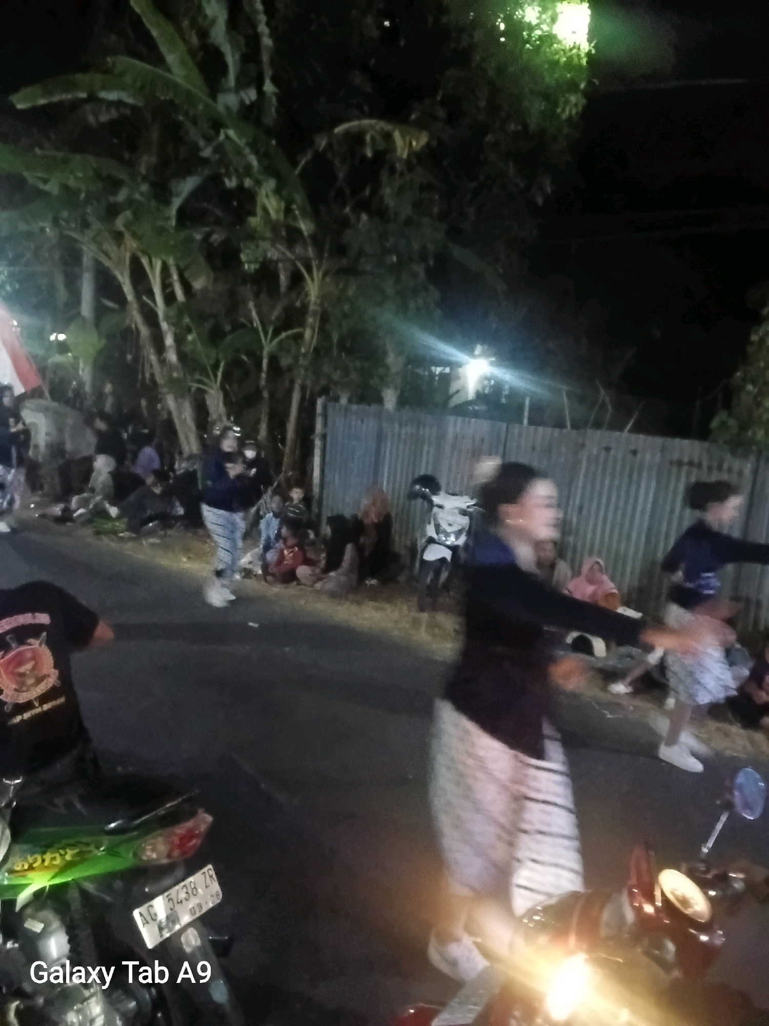This is a nighttime, eye-level, medium shot of a street scene in Wonoanti, Indonesia. A group of people, some of whom appear to be of Asian descent, are gathered along the side of a road. In the foreground, the back of a person in a black t-shirt with a circular emblem and the rear of a green and black motorcycle are visible. The motorcycle has a license plate with the characters "AG 5438 ZR" and "03-28". Further down the street, a white motorcycle is parked. Several individuals are standing and sitting, some with their backs to the camera. A woman in a dark top and a patterned skirt is in motion, possibly dancing or walking. The environment includes a corrugated metal fence behind the people and foliage with large leaves, likely banana trees, lining the background. Lighting appears to be artificial, with bright lights illuminating parts of the scene and casting shadows. The ground is asphalt, and there is some debris scattered along the roadside. The overall impression is of a casual outdoor gathering or event.