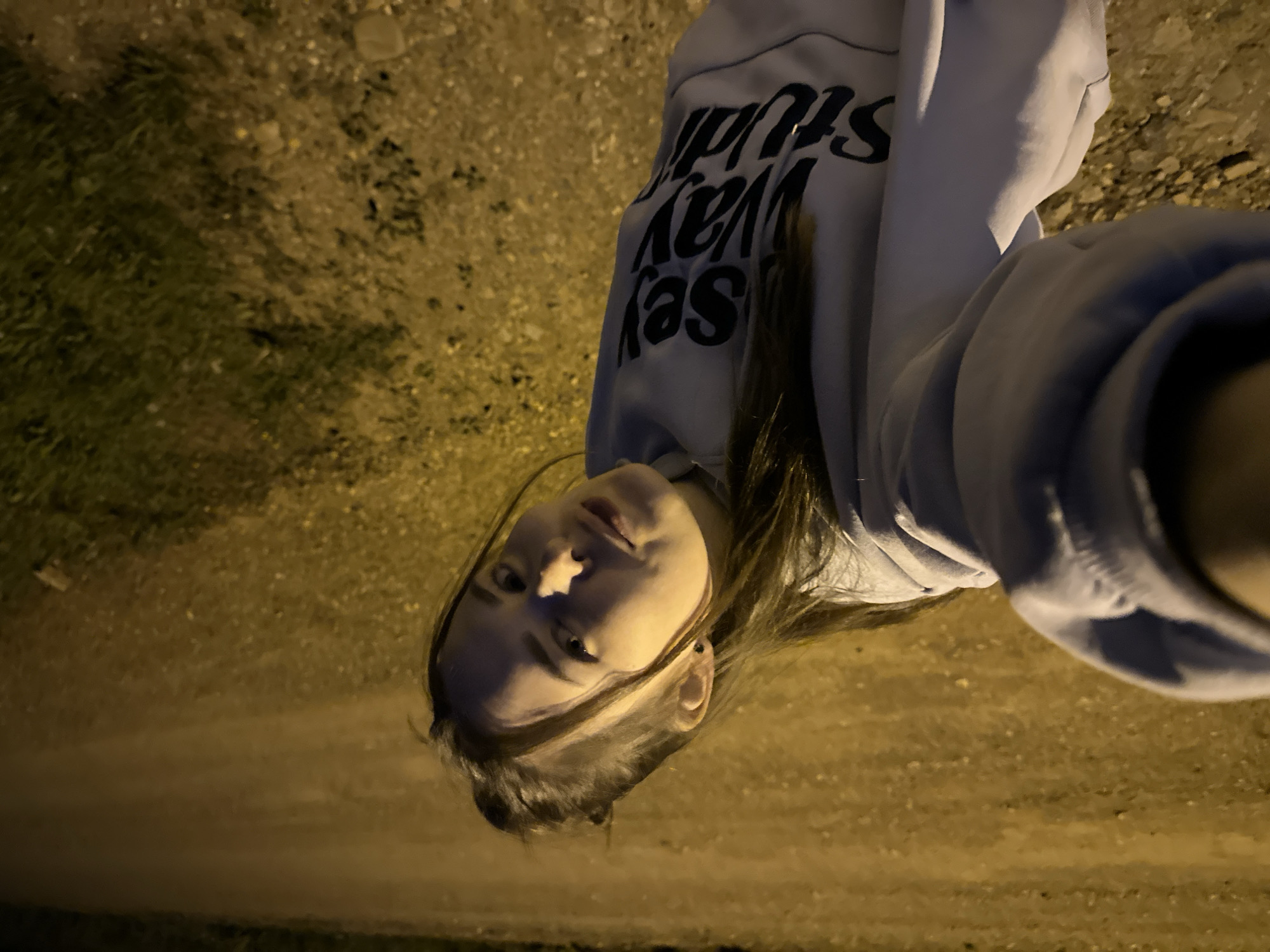 A person stands at night in what appears to be an outdoor location in Ilyinsky, Russia, based on the text on their clothing which says "Sey Way Studio" and is angled towards the camera. The environment appears to be sparsely lit, likely a pedestrian walkway or street with an uneven surface. The person is wearing a light-colored hoodie.