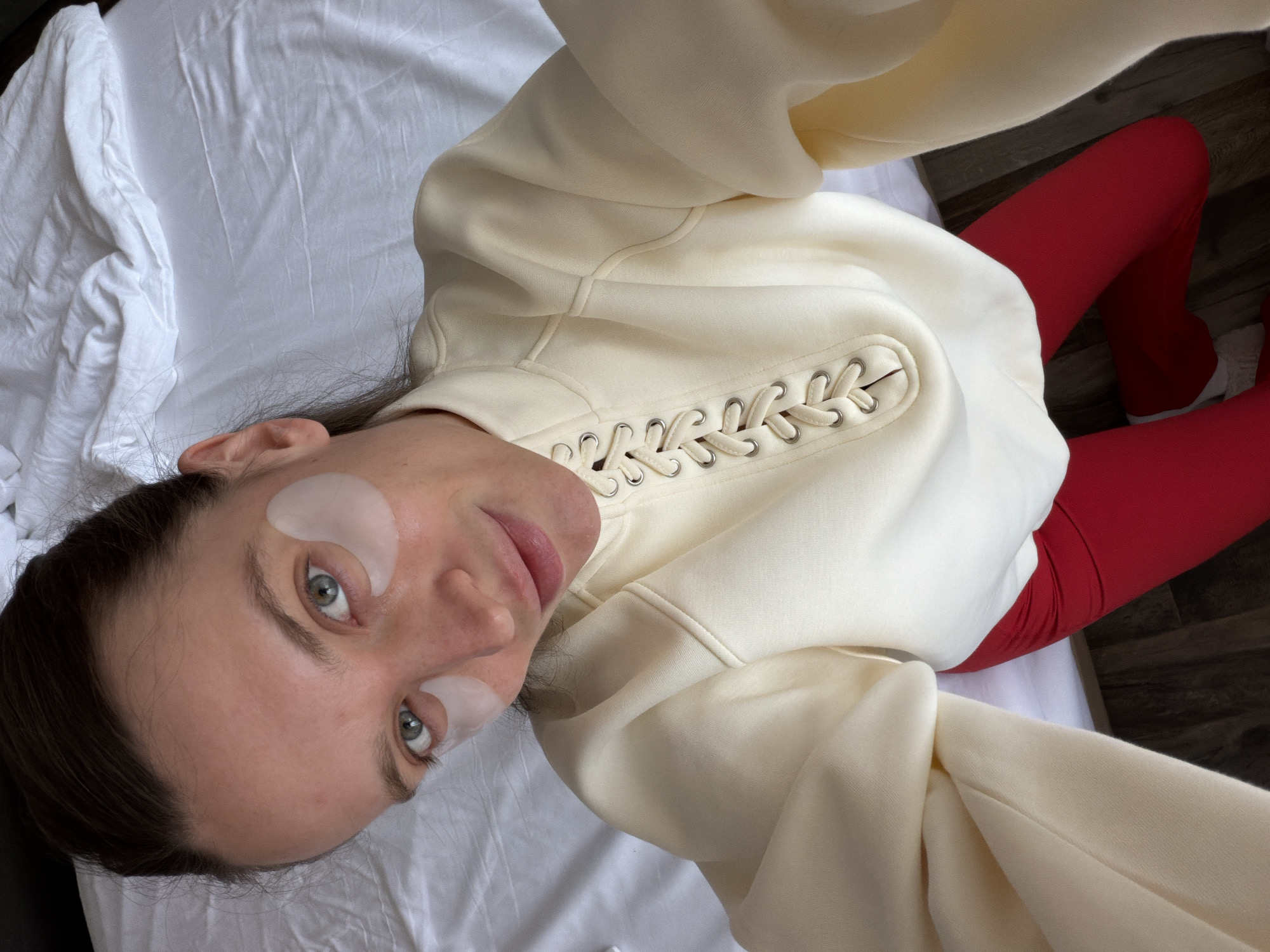 A selfie shows a woman lying on a white sheet. She is wearing eye patches, a white hooded sweatshirt with decorative lacing, and red leggings. The background includes the sheet, part of a wooden floor, and a glimpse of another white fabric. There is no information to suggest this image was taken in Budva, Montenegro.