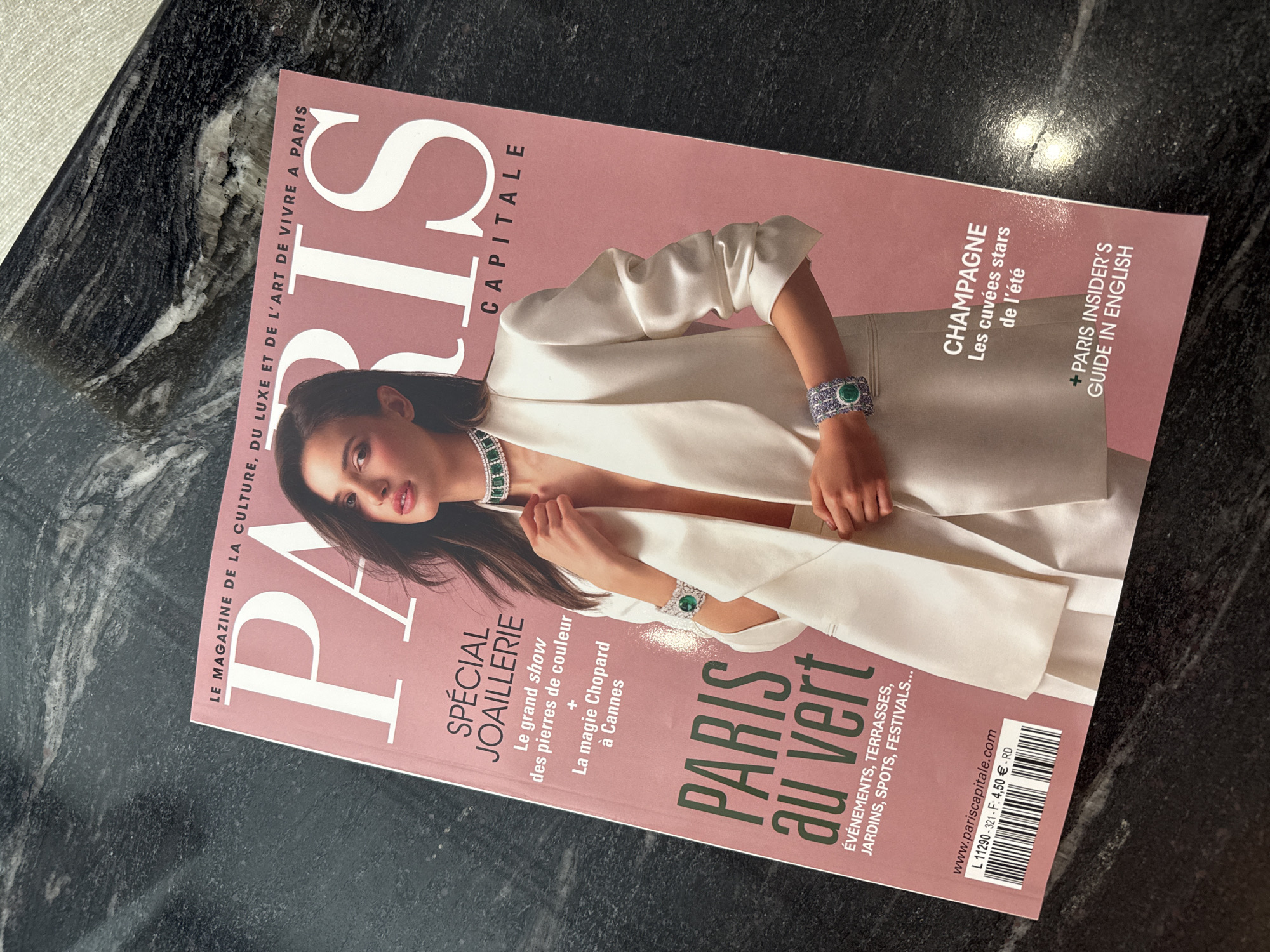 The image shows a "Paris Capitale" magazine lying on a dark, patterned surface resembling granite. The magazine cover features a woman in a white blazer adorned with emerald and diamond jewelry. The masthead "PARIS" is prominent, and there are subheadings like "SPÉCIAL JOAILLERIE," "PARIS au vert," and "CHAMPAGNE." The magazine highlights events, terraces, gardens, and festivals. It also has sections about "Paris Insider's Guide in English". The location is implied to be Paris, France, based on the magazine's title and content.