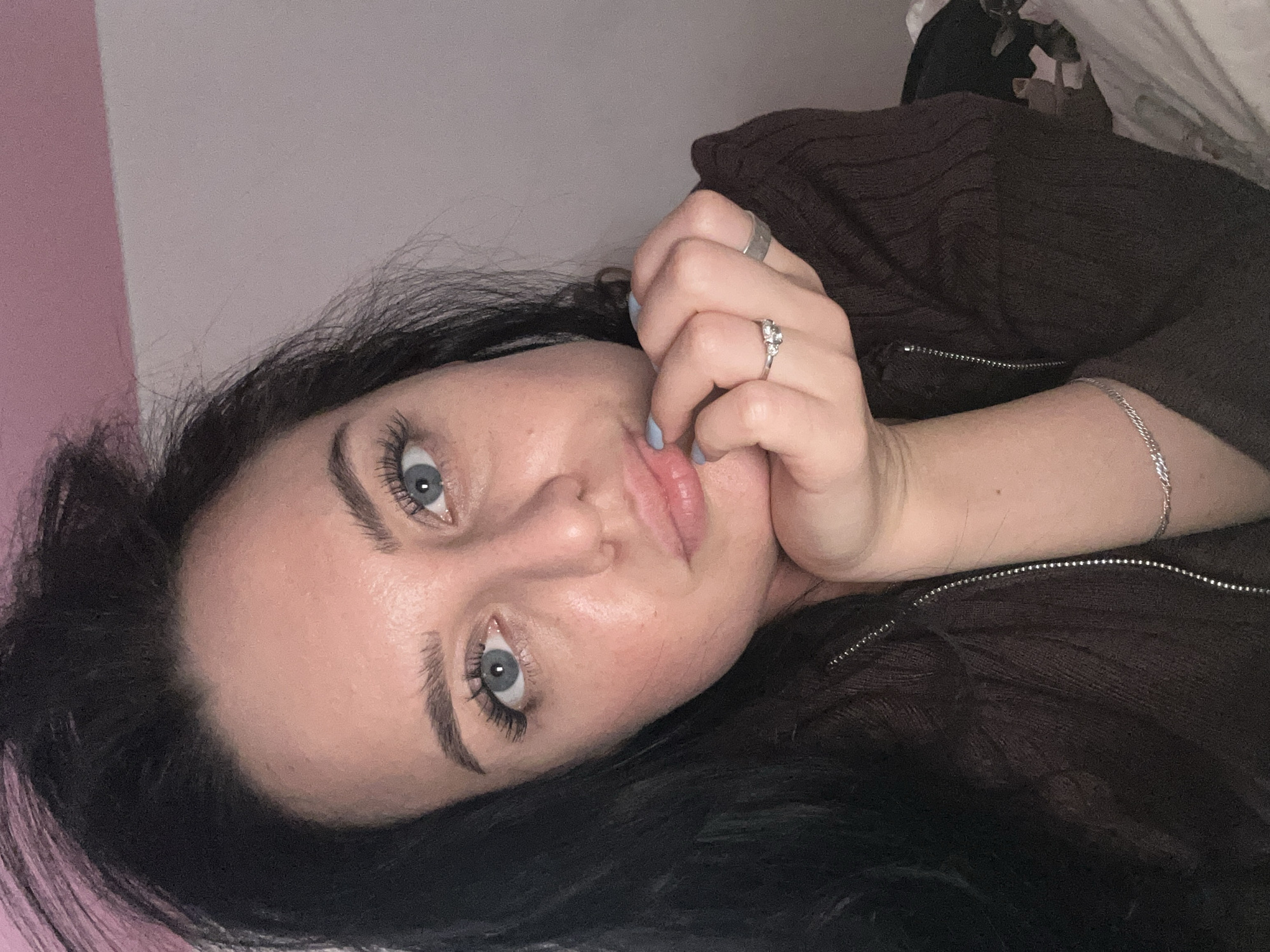 A woman with dark hair and light eyes lies on a surface with her head tilted. She is wearing a brown zip-up sweater. Her left hand, adorned with two rings, is resting on her face. A silver bracelet is visible on her left wrist. The background consists of a wall with pink and white sections. The image does not contain any information to identify the location as the city of Chaikovsky, Russia.