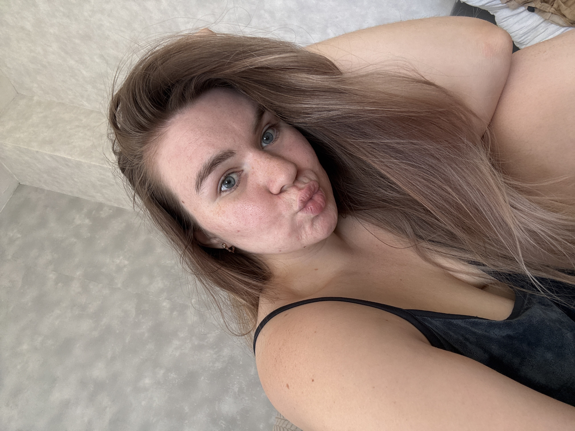 A fair-skinned woman with long, light brown hair is pictured. She is wearing a black camisole and has light blue eyes. Her lips are puckered in a kissing expression. The background appears to be a textured grey surface. The location is not discernible as Chaikovsky, Russia.