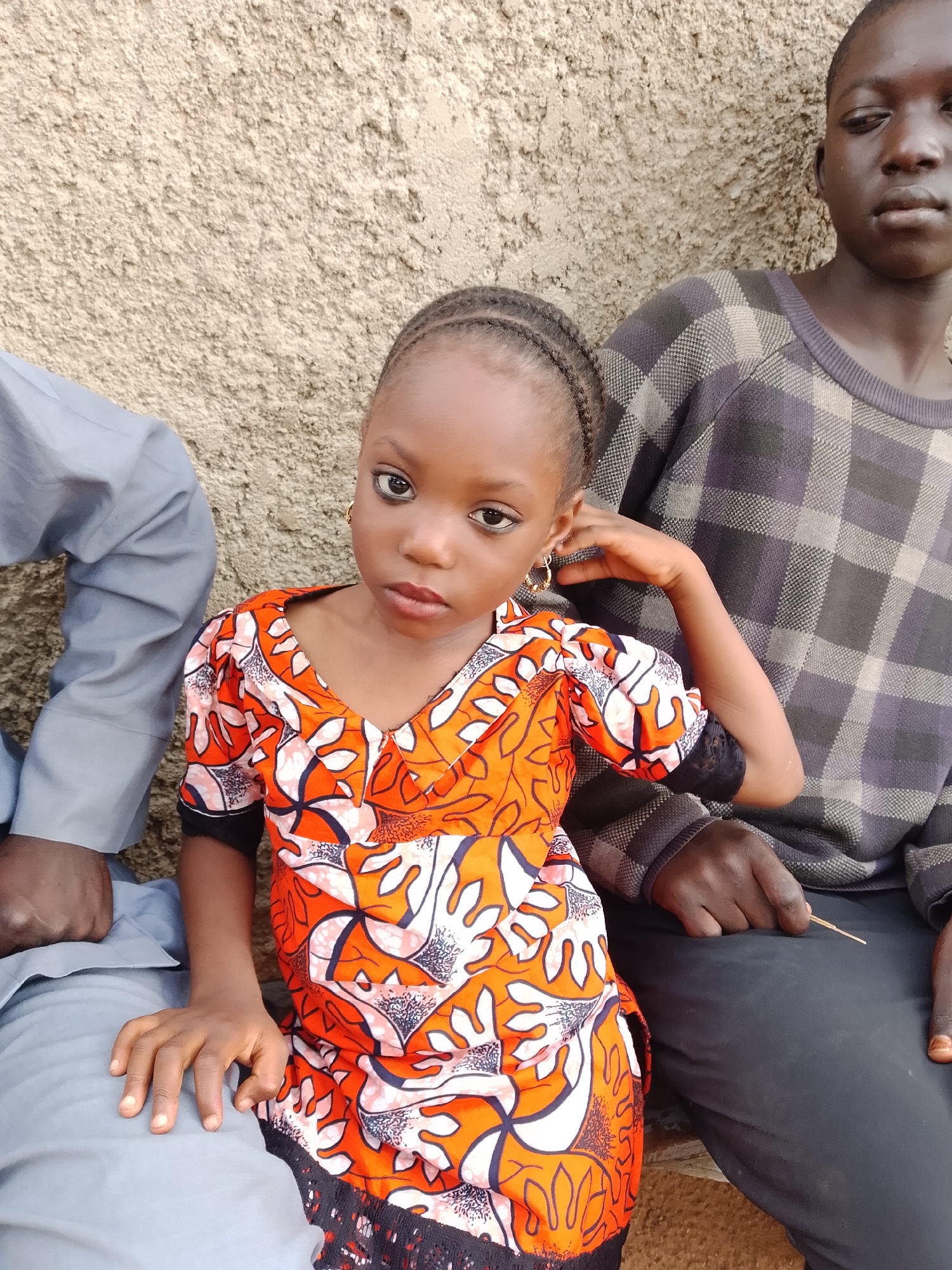 The image captures a close-up view of three individuals seated next to a textured, light-brown wall, likely in an outdoor setting. In the foreground, a young girl with dark skin and braided hair is positioned centrally, looking directly at the camera. She is wearing a vibrant orange and white patterned dress with puffed sleeves and black lace trim. To her left, a person in a light blue outfit is partially visible, only their legs and torso from the waist down are in frame. To the girl's right and slightly behind her, a young person with dark skin is visible, wearing a dark gray and black plaid sweater. The overall impression is one of a casual, possibly familial gathering. The lighting suggests it is daytime. The background wall appears to be made of concrete or stucco, common in residential or public areas in cities like Jos, Nigeria.