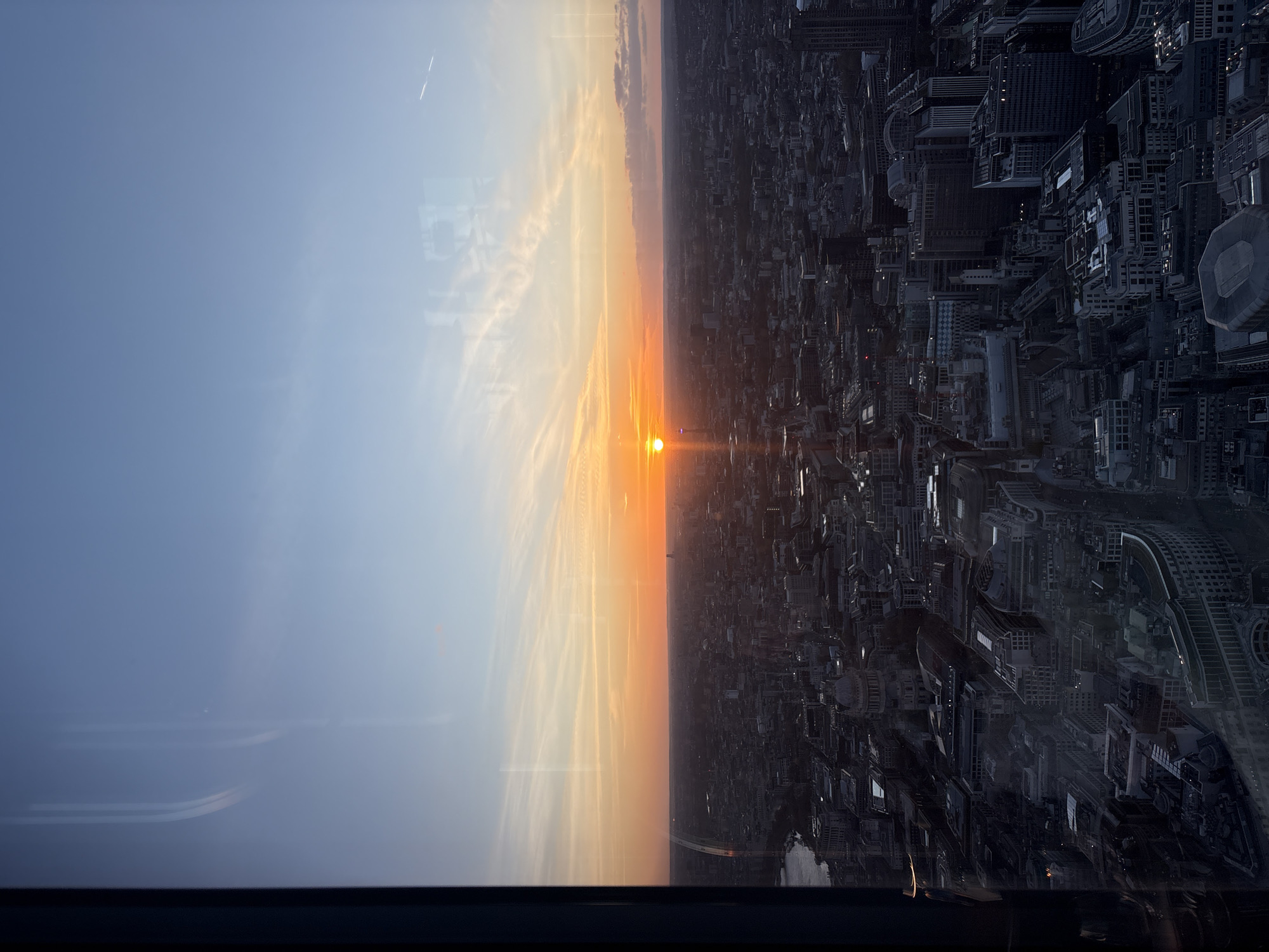 The image depicts a cityscape, most likely London, United Kingdom, observed from a high vantage point during either sunrise or sunset. On the left side of the vertical frame is a clear blue sky transitioning into an orange and yellow gradient near the center. This colored band represents the sun's position. The right side is dominated by a dense cluster of buildings and structures, typical of an urban environment. The buildings are predominantly in dark tones, offering a sharp contrast to the bright sky. A river can be seen winding through the city. There are no people visible in the image. The view is partially obstructed by a window frame at the bottom of the image.
