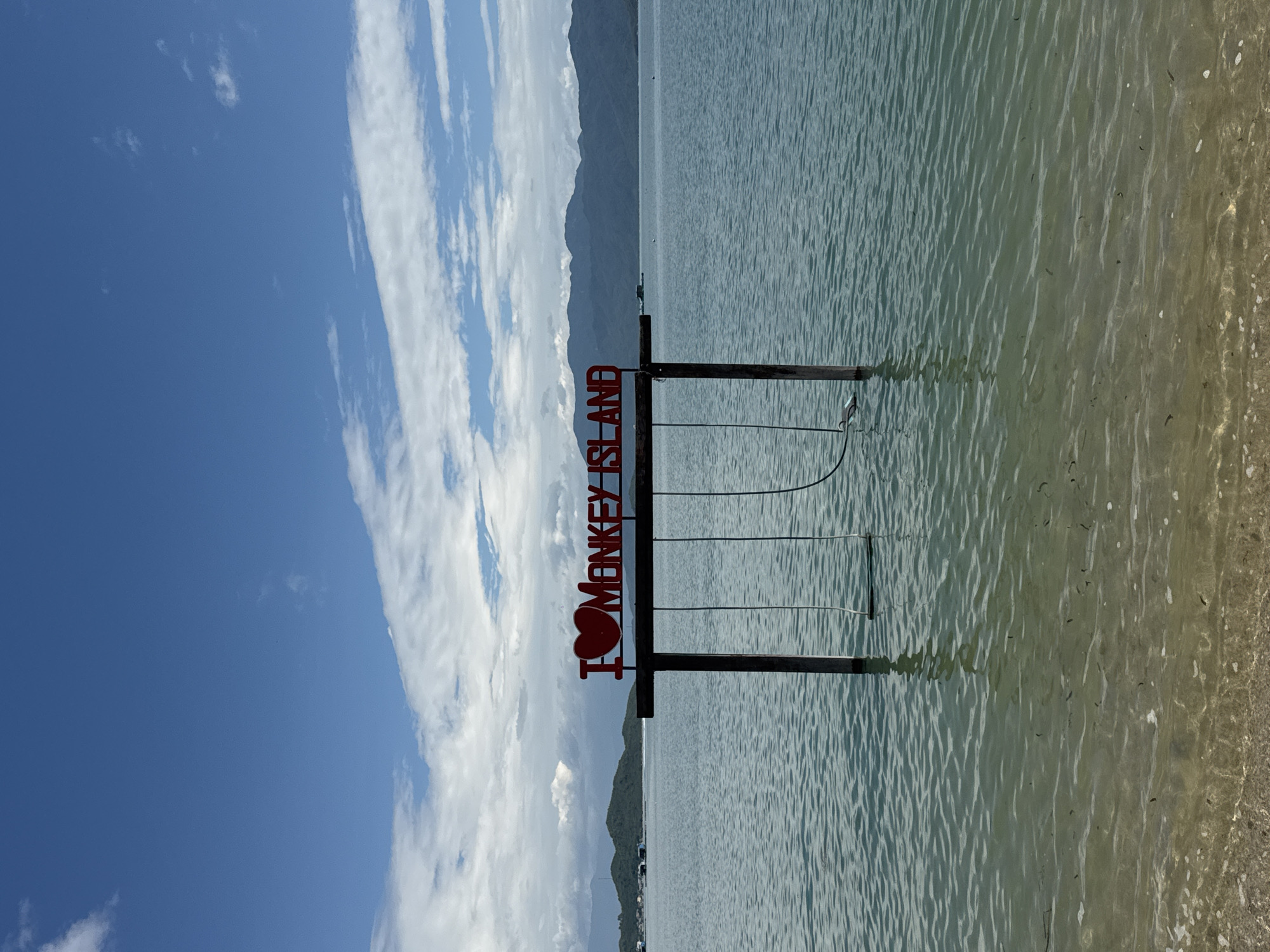 The image depicts a scenic view of Monkey Island in Ninh Ich, Vietnam. In the foreground, clear turquoise water gently laps onto a sandy shore. Two swings, suspended from a wooden frame adorned with red lettering that reads "I HEART MONKEY ISLAND," are partially submerged in the water. The wooden frame stands on posts that rise from the sea, creating an iconic photo opportunity. In the background, lush green islands dotted with distant structures and a hazy, mountainous coastline under a bright blue sky filled with scattered white clouds complete the picturesque tropical setting. The overall impression is one of a tranquil and inviting coastal destination.