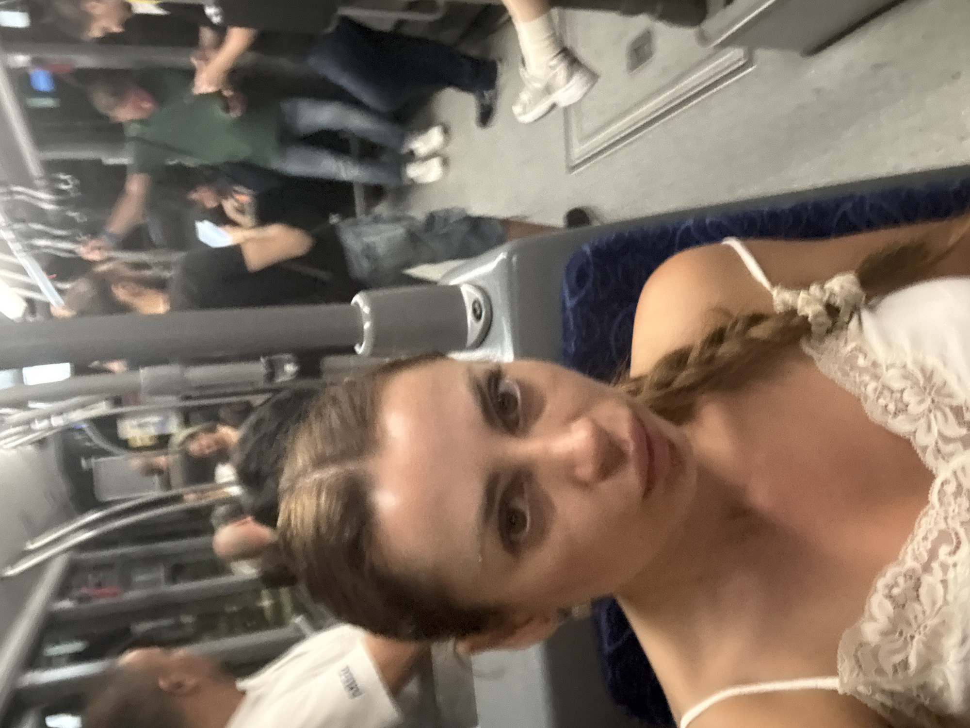 The image depicts the interior of a public transportation vehicle, likely a bus or tram, filled with passengers. A woman in the foreground is taking a selfie. She has long, braided brown hair and is wearing a white lace-trimmed camisole. In the background, several other individuals are visible, some standing and holding onto overhead rails, while others are seated. The overall lighting suggests an indoor environment, possibly during the day or evening. The image does not contain any specific visual cues to indicate the city of Venice, Italy.