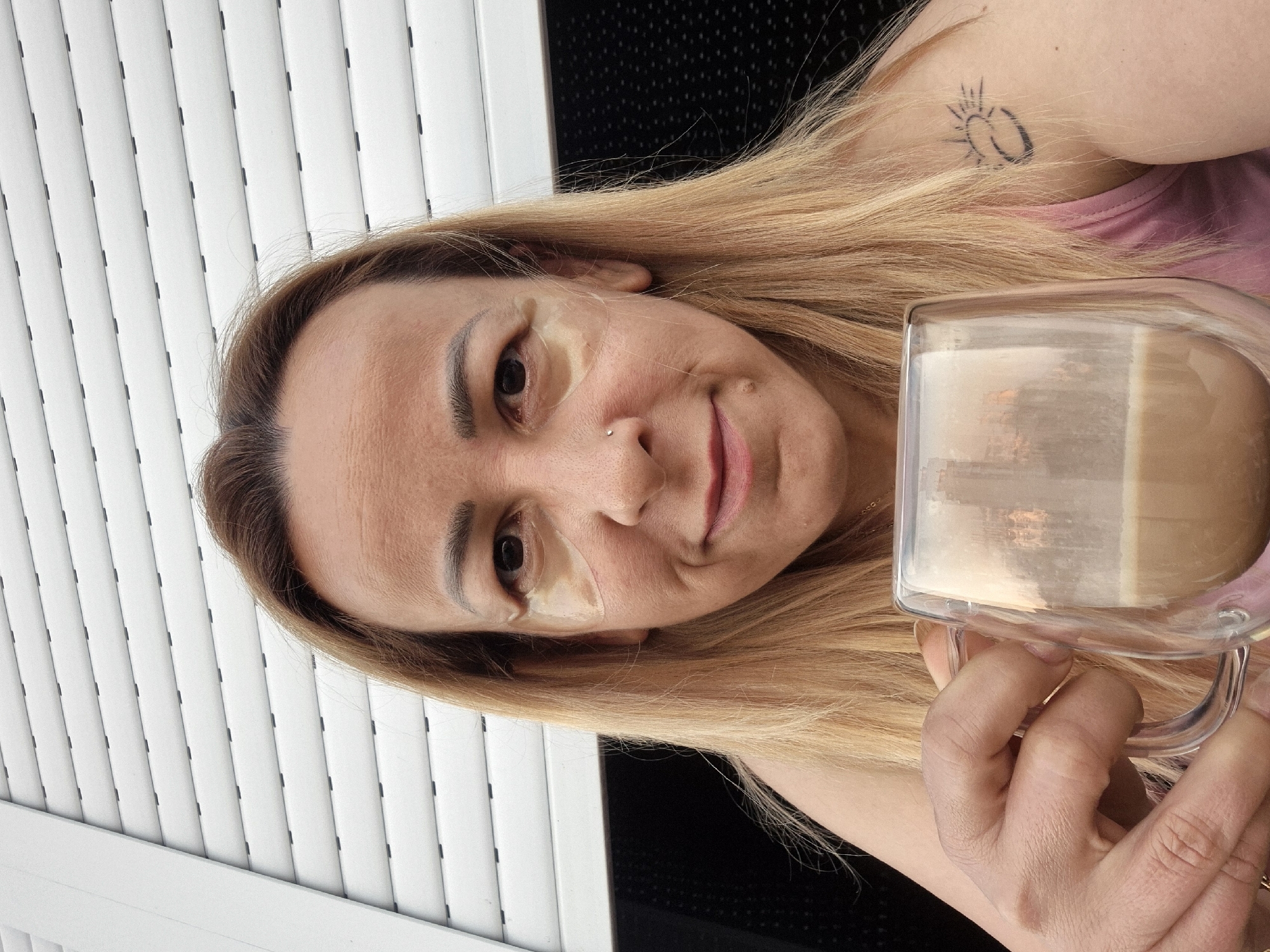 A close-up, waist-up, outdoor, selfie-style portrait shows a blonde woman with light skin, who is holding a clear, double-walled glass mug of coffee in her right hand. She is wearing a pink tank top and has dark blonde hair. She has eye patches under her eyes and a nose ring. A small tattoo of a sun is visible on her left shoulder. The background consists of white horizontal blinds and a black textured surface. The woman is looking directly at the camera and smiling slightly. The lighting suggests it is daytime, with some reflections visible in the coffee mug. The location is unknown.