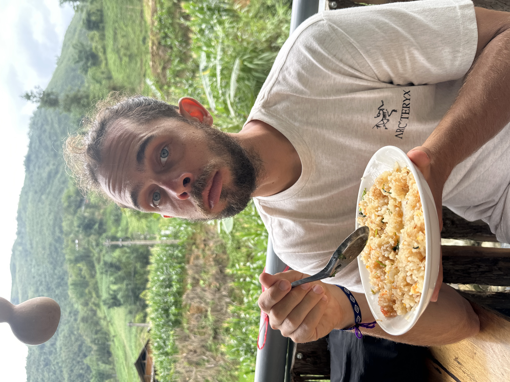A man with light skin, a beard, and blue eyes is holding a plate of fried rice with a spoon, posing in front of a background of lush greenery that includes mountains. The man wears a light-colored T-shirt with the "Arc'teryx" logo. He is wearing a dark-colored garment from the waist down, and a bracelet on his left wrist. The outdoor setting suggests the image was taken in Thanh Bình, Vietnam, although the exact location is not discernible.