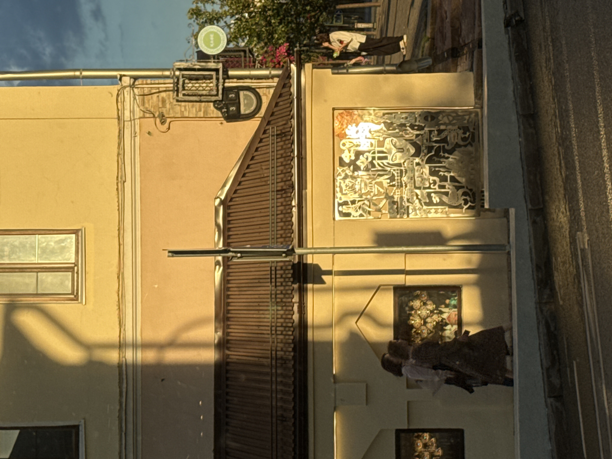 The image shows a street scene in Kazan, Russia, during sunset. A yellow building dominates the frame, with a corrugated metal roof partially visible. Two women are walking on the sidewalk in front of the building. One is wearing a patterned dress, and the other is wearing a white shirt and dark pants. They are walking towards the left side of the frame. A large, abstract mural is displayed on the wall of the building, featuring stylized faces and figures. To the right, a person in dark pants and a white shirt stands near a sign that reads "KAFE." Trees with pink flowers are visible in the background. The setting sun casts long shadows across the scene, illuminating the building and sidewalk with a warm, golden light.