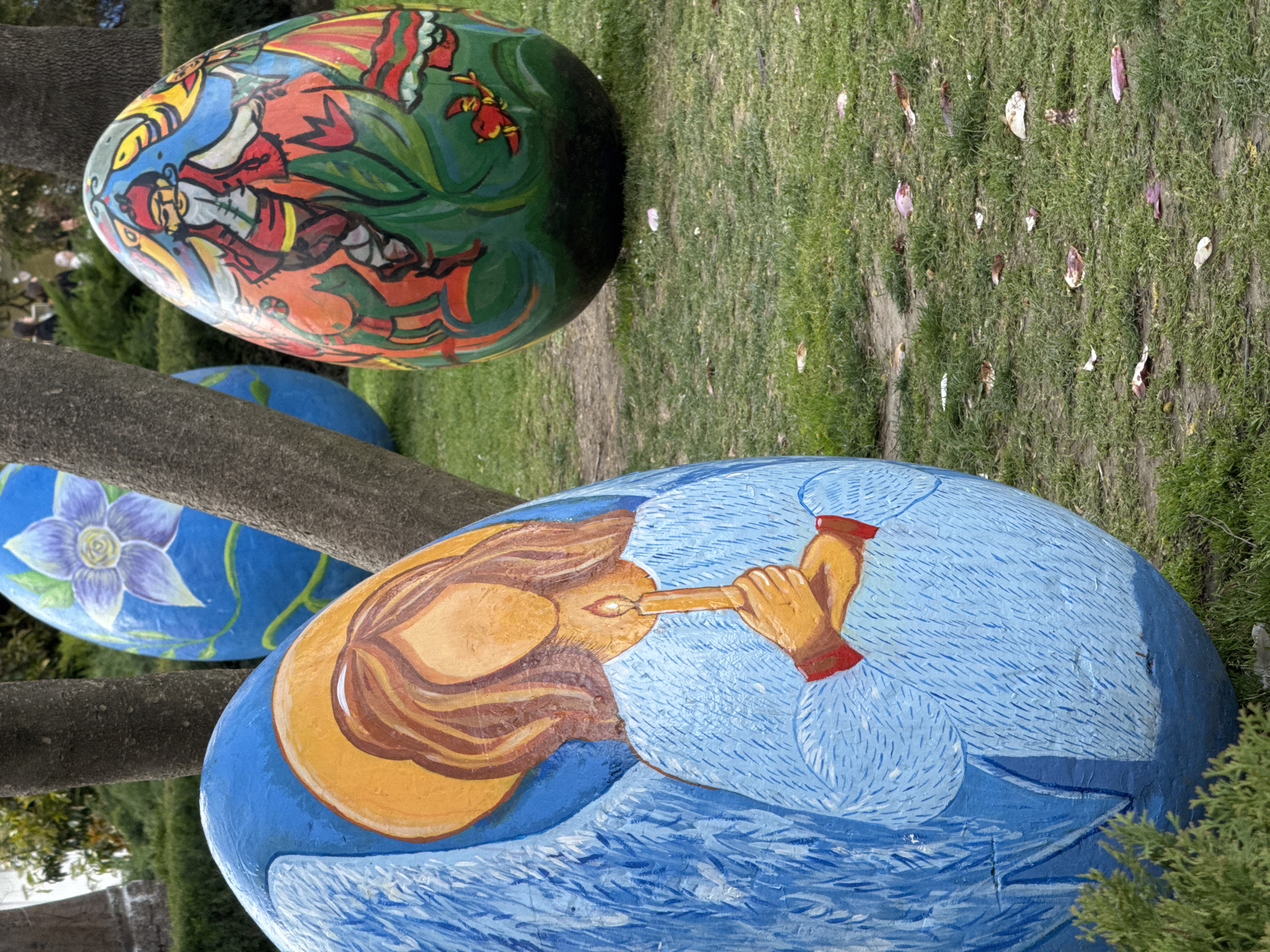 The image shows three large, decorated Easter eggs displayed in a park in Plovdiv, Bulgaria. The eggs are resting on grass dotted with fallen petals.
The first egg is green and features a painted figure in red and white clothing surrounded by green foliage. The second egg is blue and painted with a purple flower. The third and closest egg is also blue, featuring a person with tan skin and brown hair playing a long, thin instrument. The person's hands are visible, with red accents at the wrists.
The eggs are positioned among tree trunks, with the background showing a grassy area and scattered leaves.