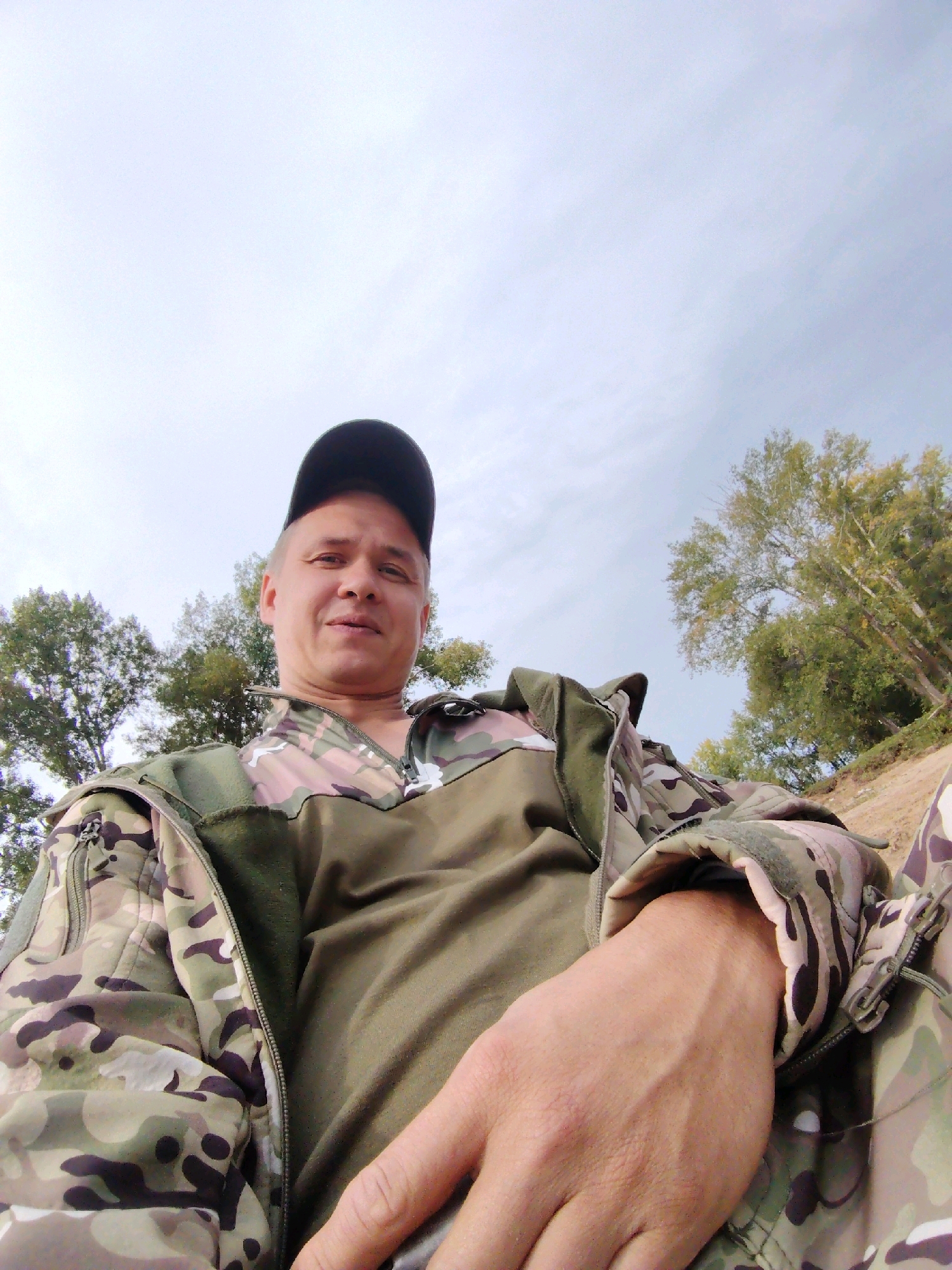 In Noviy, Russia, a low-angle, outdoor, selfie-style photograph shows a man wearing a camouflage jacket and a black baseball cap. He is looking towards the camera with a neutral expression. The background features a sky with some clouds, trees, and a sandy embankment. The man's right arm and hand are prominently in the foreground, resting on his lap.