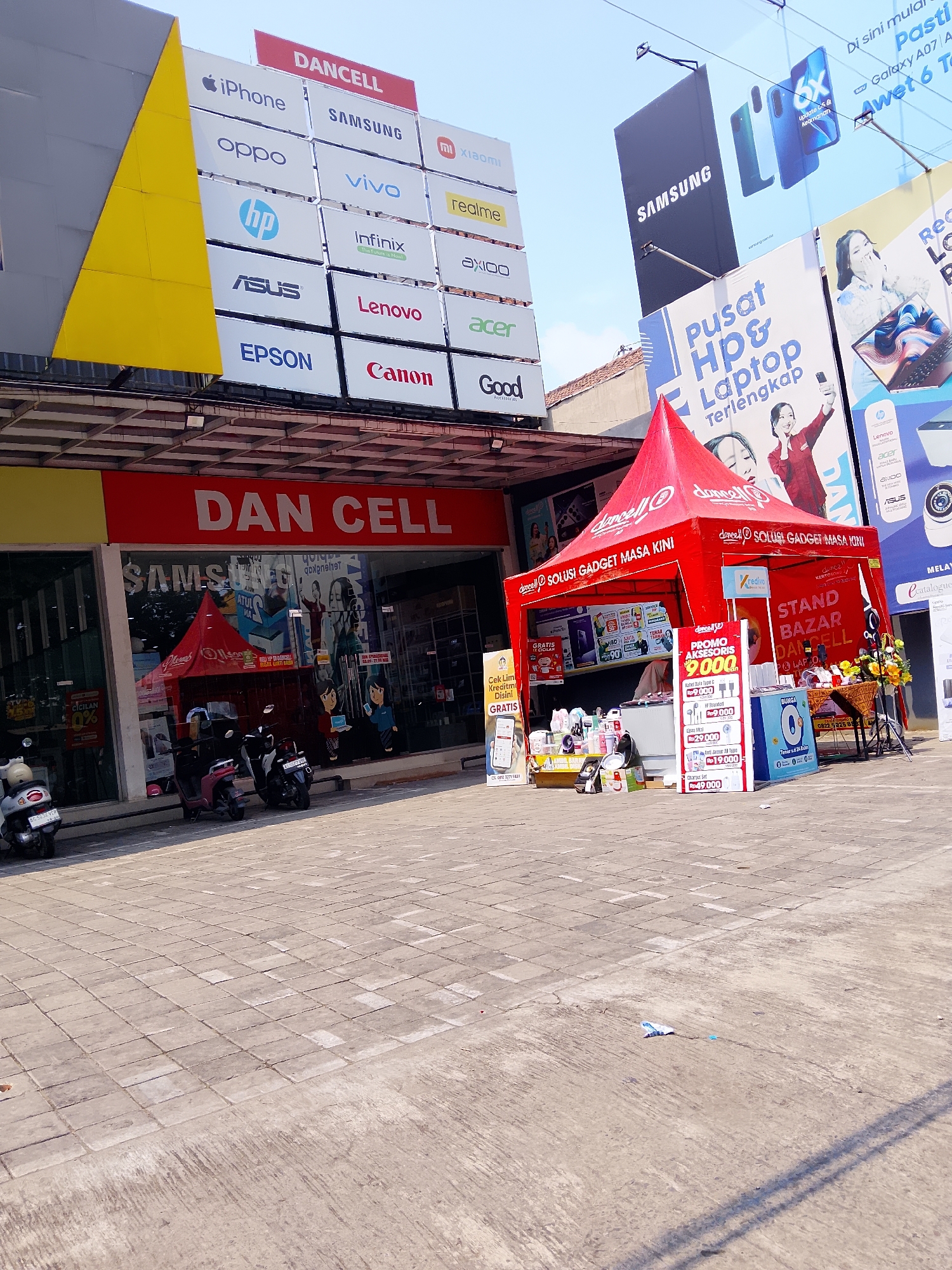 This is an exterior, eye-level, medium shot of a Dan Cell electronics store in Kertosono, Indonesia, on a bright, sunny day. The store's facade is prominent, with large red lettering spelling out "DAN CELL." Above the store, a series of illuminated panels display various electronics brands like iPhone, Samsung, Oppo, Vivo, Infinix, Lenovo, Canon, and Epson. To the right of the store, a red pop-up tent is set up, serving as a promotional stand for "Dan Cell Solusi Gadget Masa Kini" (Dan Cell Modern Gadget Solutions). This tent displays various electronics and accessories, with pricing information visible on signs, including "PROMO AKSESORIS 9.000" and other items priced at Rp 29,000 and Rp 49,000. A sign on the tent reads "STAND BAZAR DANCELL LAPTOP." In front of the store and tent, the paved ground is a mix of square tiles and concrete, with several parked scooters and motorcycles visible. Further to the right and in the background, large billboards advertise Samsung phones and other electronic products, including an image of a Samsung phone and text in Indonesian promoting "Pusat HP & Laptop Terlengkap" (Complete HP & Laptop Center). The overall environment suggests a busy commercial area focused on electronics retail.