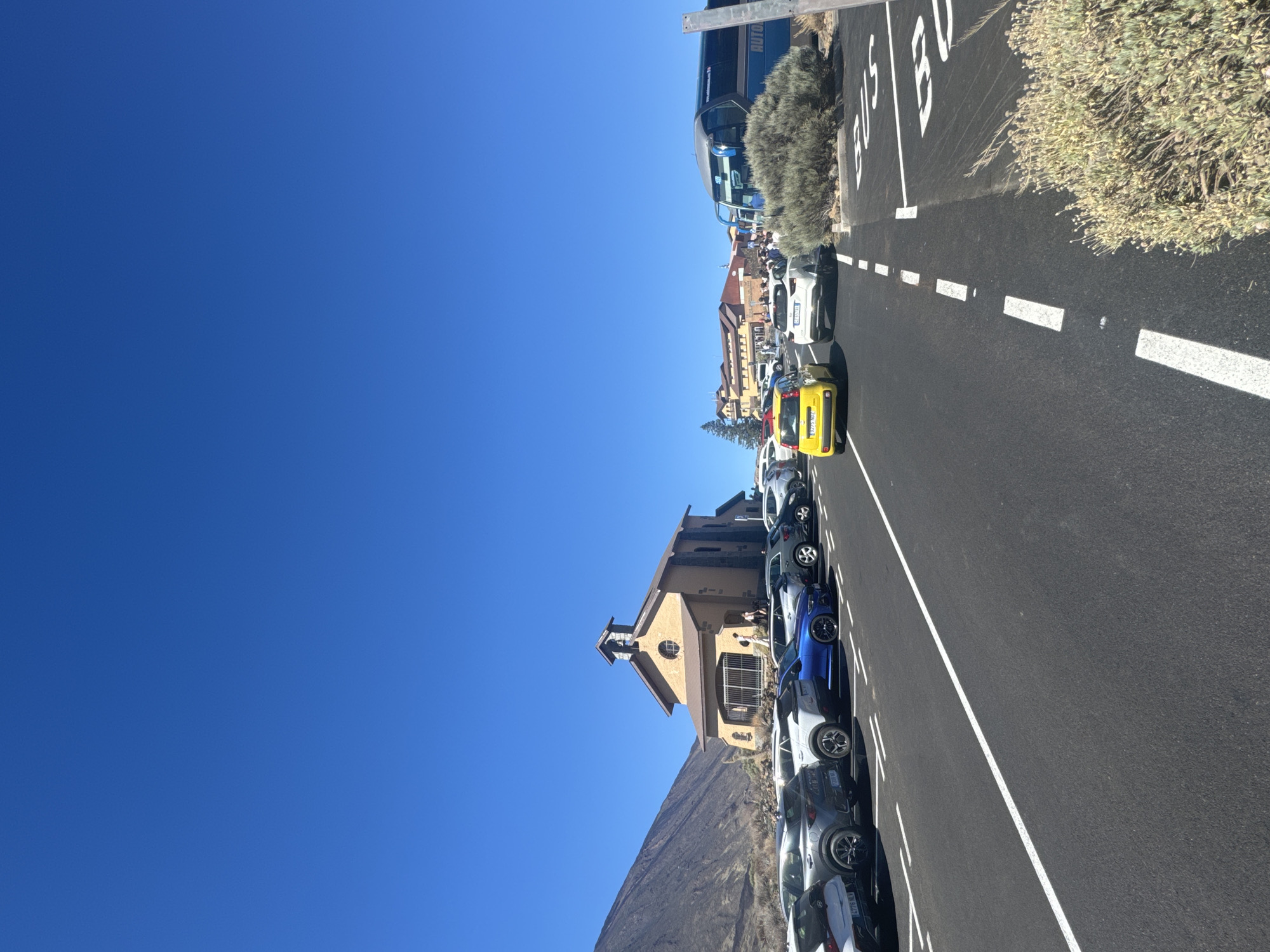 The image shows a parking lot with several cars and a bus under a clear blue sky. In the background, there is a small church-like building with a bell tower, and a mountain slope to its left. Further in the background, more buildings are visible, suggesting a populated area. A blue bus is parked on the right side of the frame, with the word "AUTO" visible on its side. The parking lot has white lane markings and some sparse vegetation on the sides. People are seen walking near the buildings and the bus. This scene is located at the Roques de Garcia, a geological formation in Teide National Park, which is part of the municipality of La Orotava, Spain.