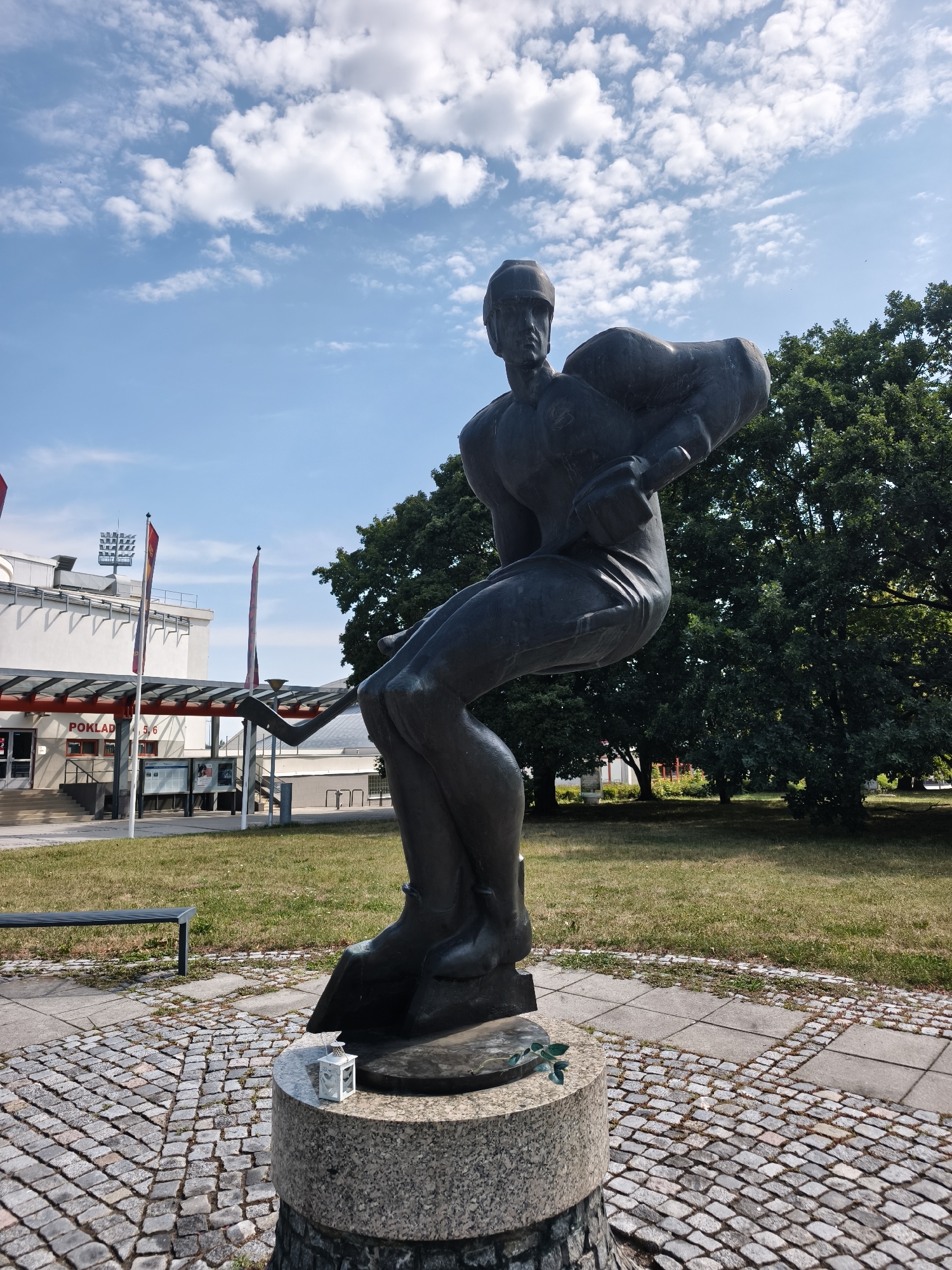 A bronze sculpture of a male ice hockey player in mid-action is positioned in front of a building with a banner reading "POKLADNA" (ticket office). The sculpture is on a stone pedestal, which has a small white lantern and a green leaf decoration. The sculpture is dark grey and depicts a player wearing a helmet and holding a hockey stick. The background features a grassy area, trees, and a blue sky with white clouds. Flags on poles are visible near the building entrance. The paving around the sculpture is made of cobblestones. The sculpture is located in Pardubice, Czechia, outside of the Enteria Arena, which is home to the Dynamo Pardubice ice hockey team.
