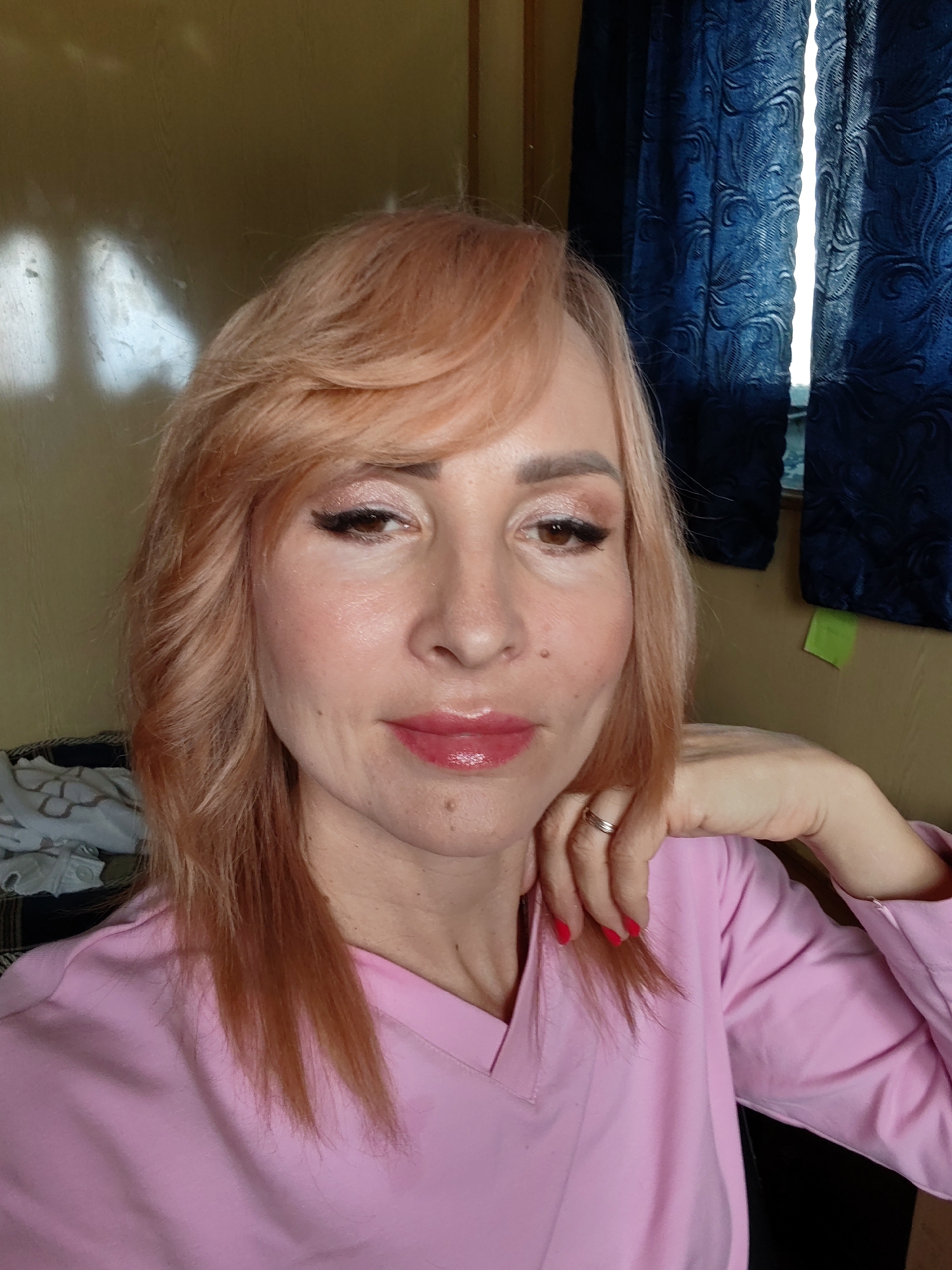 A medium shot image captures a woman with light-colored hair, wearing a pink V-neck shirt. Her right hand rests on her chin and her left arm is bent with her hand near her neck. She is looking at the camera with makeup on her eyes and lips. She is wearing a ring on her left hand. The background is a plain yellow wall with blue curtains and a window to the right. The woman is indoors. The image is a selfie.