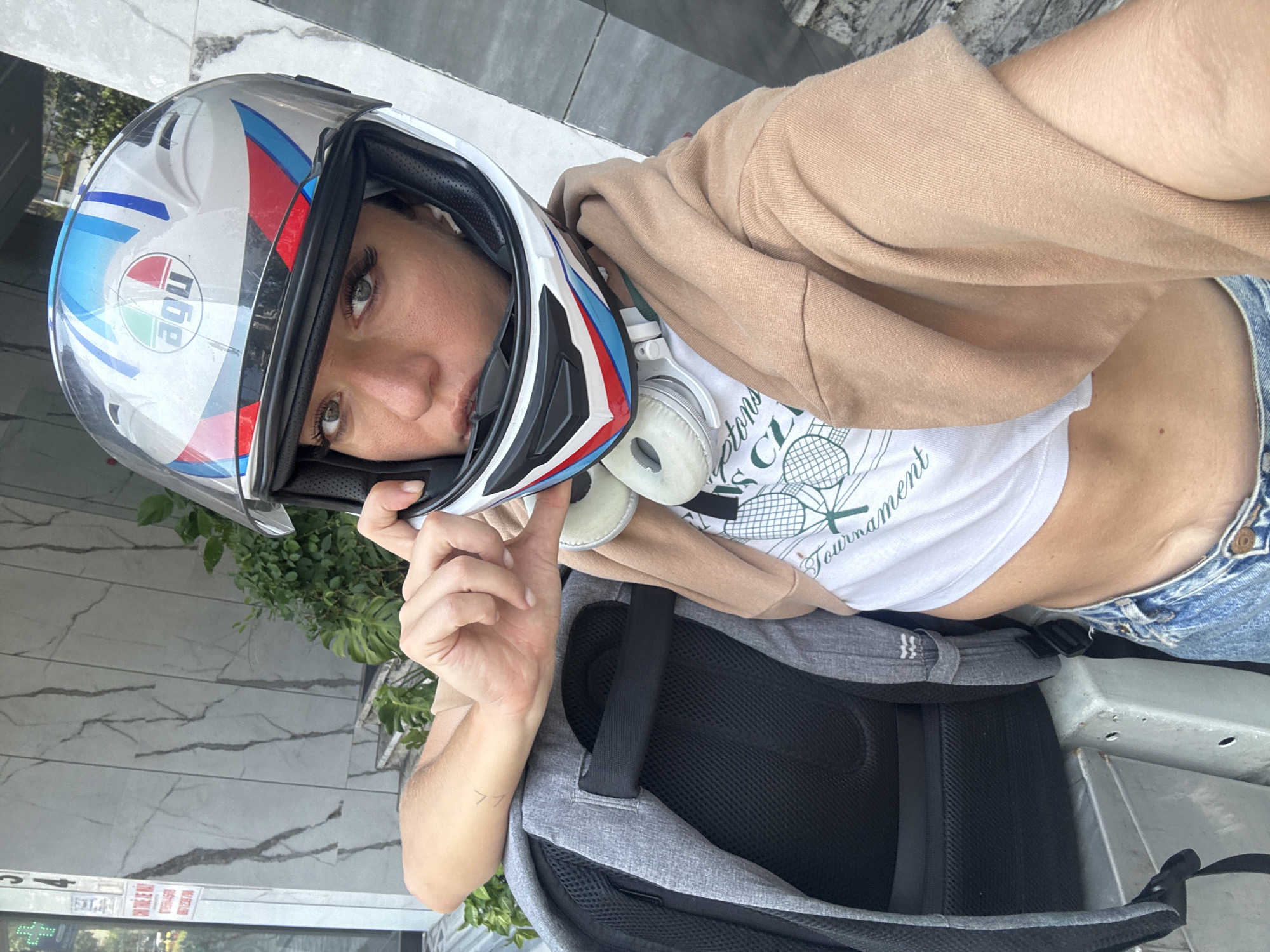 A person with fair skin, wearing an AGV motorcycle helmet, a tan hooded sweatshirt, a white crop top t-shirt with a green and white graphic, and blue denim shorts, is posing for a photo. The person is wearing white headphones around their neck and has a grey and black backpack. The background shows a building with marble-like paneling and greenery. The photo was taken outside of a pharmacy store on Trần Cao Vân Street, in Đà Nẵng, Vietnam.