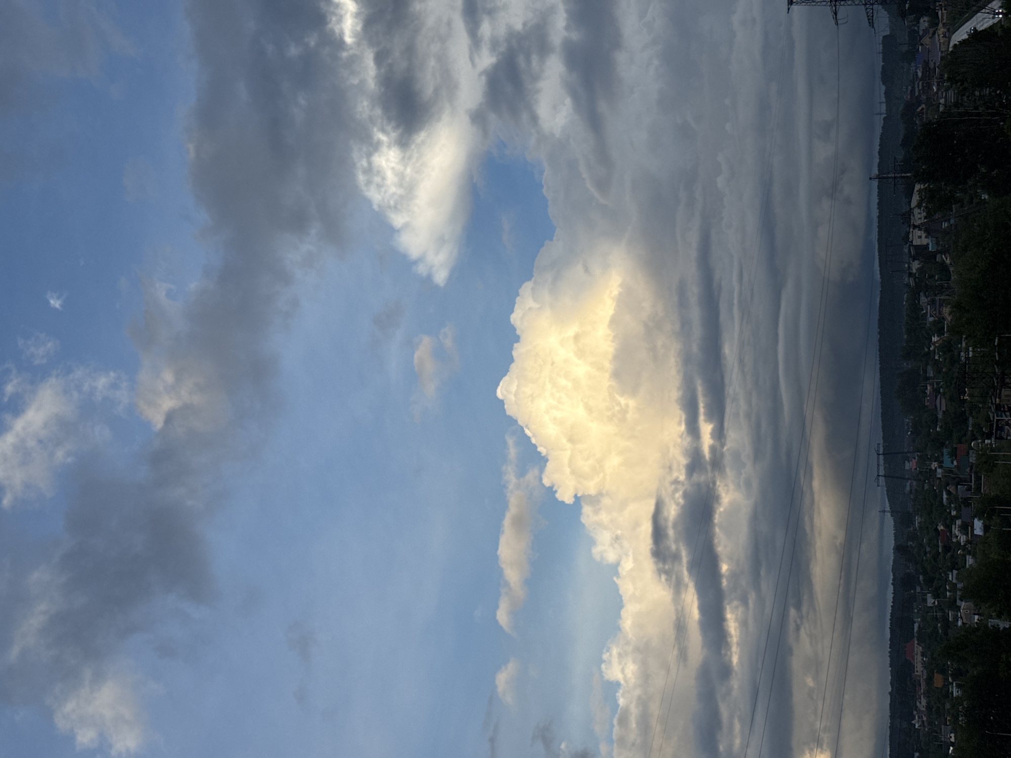 The image displays a vast sky dominating the frame, characterized by a dramatic interplay of light and shadow across various cloud formations. The upper left portion of the sky is a clear, light blue, with scattered, wispy white clouds. Moving towards the center and right, large, dense cumulus clouds are prominent, some brilliantly illuminated with a golden-yellow hue by the sun, suggesting late afternoon or early evening light. Other clouds, particularly towards the right side and lower parts of the sky, are dark grey and layered, indicating a heavier cloud cover or approaching weather.
Beneath the extensive sky, a distant landscape stretches across the bottom quarter of the image. This landscape features a dark, forested area intermingled with numerous small buildings, likely houses, indicating a town or village situated along a body of water. A long, dark horizontal band suggests a river or a large lake running through the scene. Several tall utility poles with power lines are visible, cutting across the sky and landscape, adding an element of human infrastructure to the natural setting. No people or visible text are present in the image. The overall impression is one of a vast, dynamic sky over a serene, populated natural landscape.