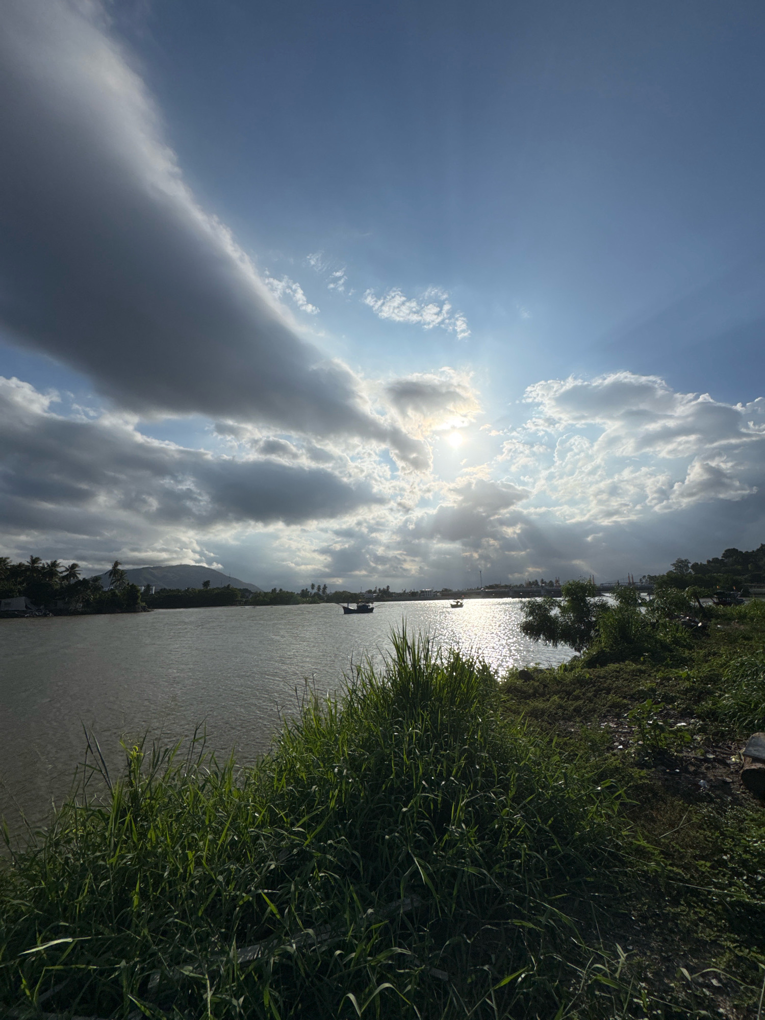 A wide, calm river flows through a lush landscape under a dramatic sky. In the foreground, tall, green grass grows along the bank. The river's surface reflects the bright sunlight, creating shimmering patterns. In the distance, a line of trees and hills stretches across the horizon. Two small boats are visible on the water, one closer to the left bank and another further out. The sky is a mix of deep blue and heavy, grey clouds, with rays of sunlight piercing through, creating a dramatic contrast. The scene appears to be a serene, natural setting on the outskirts of Nha Trang, Vietnam.