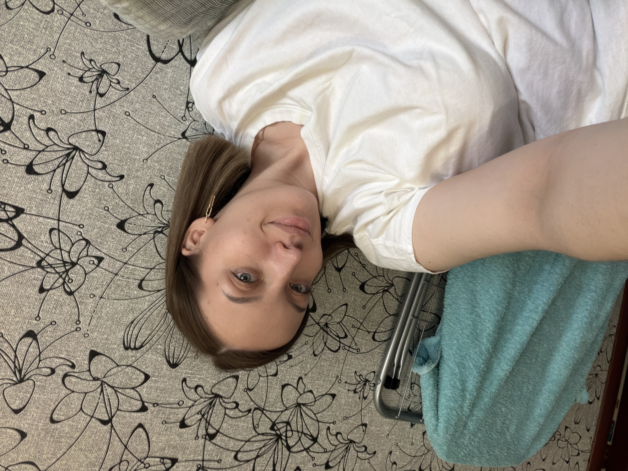 A woman with light brown hair and fair skin, wearing a white T-shirt and gold earring, lies on a patterned gray surface with black floral designs. A light blue towel is draped over a metal frame to her right. The location cannot be determined from the image.