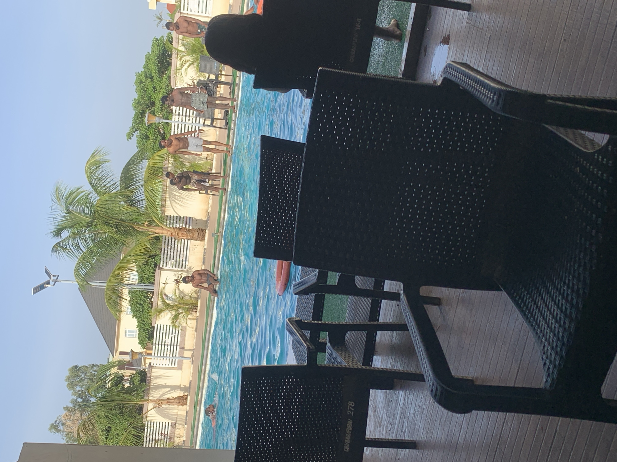 The image shows a swimming pool scene in what appears to be Barakin Lerawi, Nigeria. A group of men are standing near the edge of the pool. A palm tree is visible near the pool. In the foreground are dark-colored chairs, obscuring the view partially. A person is seated in one of the chairs. Someone is jumping into the pool while another is in the pool. A solar light is also visible, along with a building. The scene is shot in daylight.