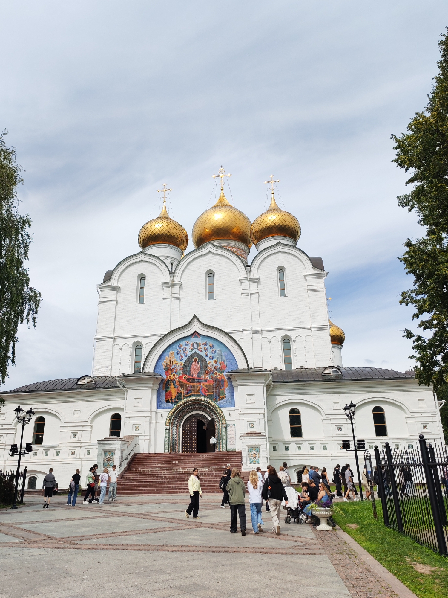 The image captures a wide-angle view of the Assumption Cathedral in Rusanovo, Russia, under a cloudy sky. The cathedral is a white, multi-domed structure adorned with gold-colored onion domes topped with crosses. A large mosaic depicting a religious scene is prominently displayed above the main entrance. A wide set of stairs leads up to the entrance. In front of the cathedral, a paved area is frequented by a diverse group of people. Some individuals are walking, while others are standing or sitting. There's a black metal fence lining the right side of the scene, separating the paved area from a grassy strip and trees. The overall impression is that of a public space around a significant religious landmark on a day with mixed weather conditions.