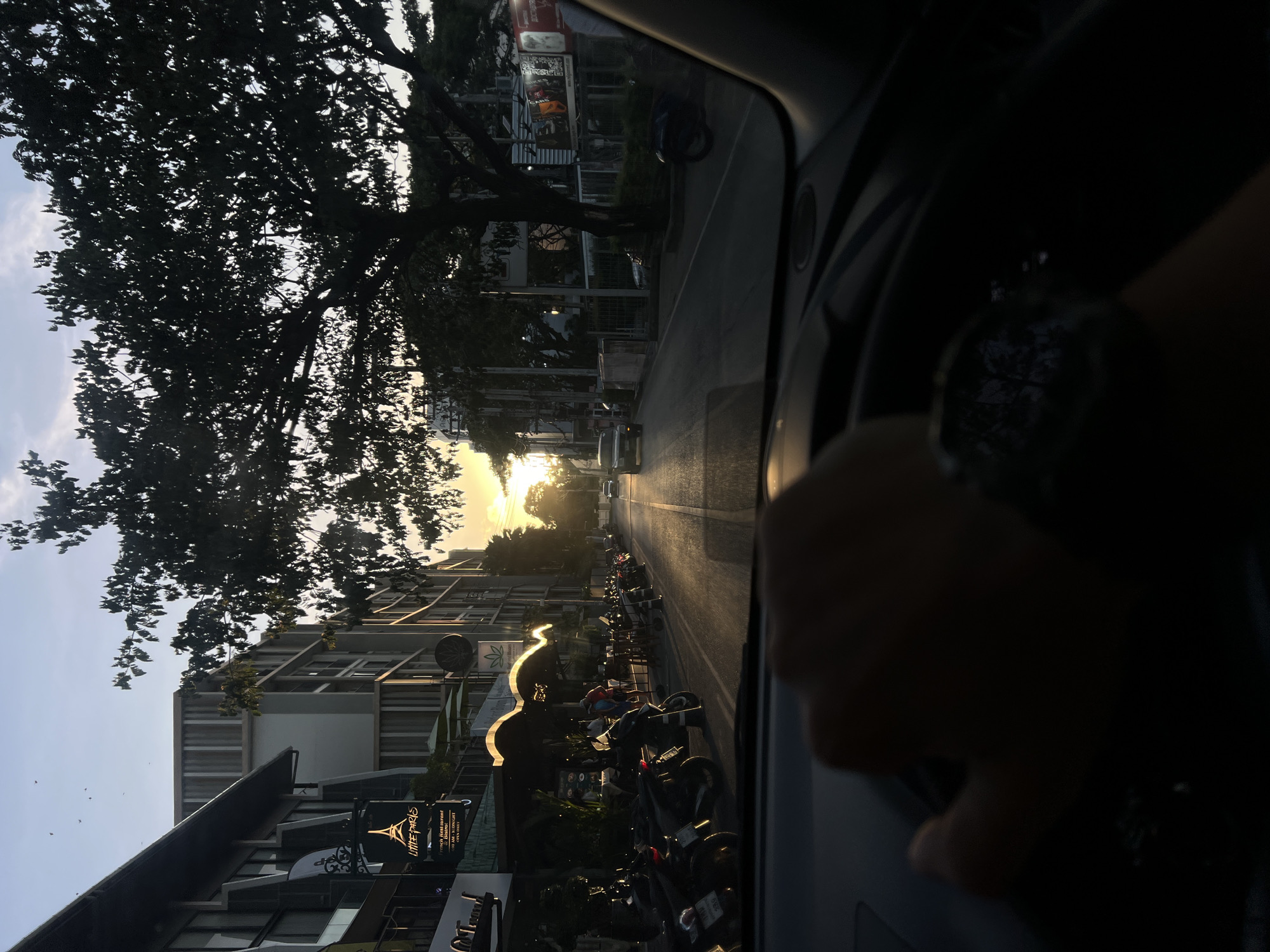 The media file is an image taken from inside a car, looking out through the windshield. The perspective is rotated, making it appear as though the car is driving on a wall. The scene depicts a street lined with buildings and trees, with a row of motorcycles parked along the side of the road. The sun is setting, casting a warm glow on the street and buildings, and silhouetting the trees. There is a person's hand visible in the foreground, resting on the steering wheel.
The subjects are the street, buildings, parked motorcycles, trees, and the setting sun. The scene suggests a typical urban or suburban environment. The activity is driving, implied by the point of view from inside a car and the hand on the steering wheel. The time of day is clearly sunset due to the golden light and long shadows. The weather appears clear, with a bright sky.
Notable details include the stark contrast between the silhouetted trees and the bright sky, the reflections of the sunset on the wet road surface, and the various architectural styles of the buildings. One building has a sign that reads "Little Paris" with an Eiffel Tower illustration, suggesting a French restaurant or cafe. Another sign beneath it appears to advertise a "French restaurant & Bistro". The overall mood is peaceful and serene, characteristic of a sunset drive.