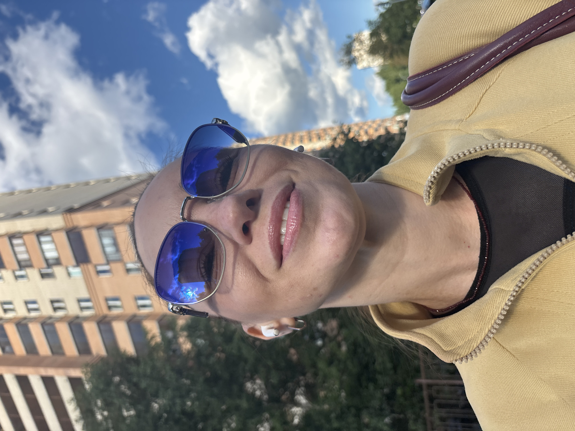 A close-up, low-angle selfie shows a woman wearing sunglasses, a yellow zip-up jacket over a black mesh shirt, and a thin beaded necklace. She has white wireless earbuds in her left ear and a small hoop earring. Her lips are glossy and slightly parted. Her reflection in the sunglasses shows the blue sky with white clouds and a building. In the background, a tall, multi-story building with a tan facade and dark windows is visible, along with green trees and a bright blue sky with scattered clouds. The location is identified as a city in Russia.