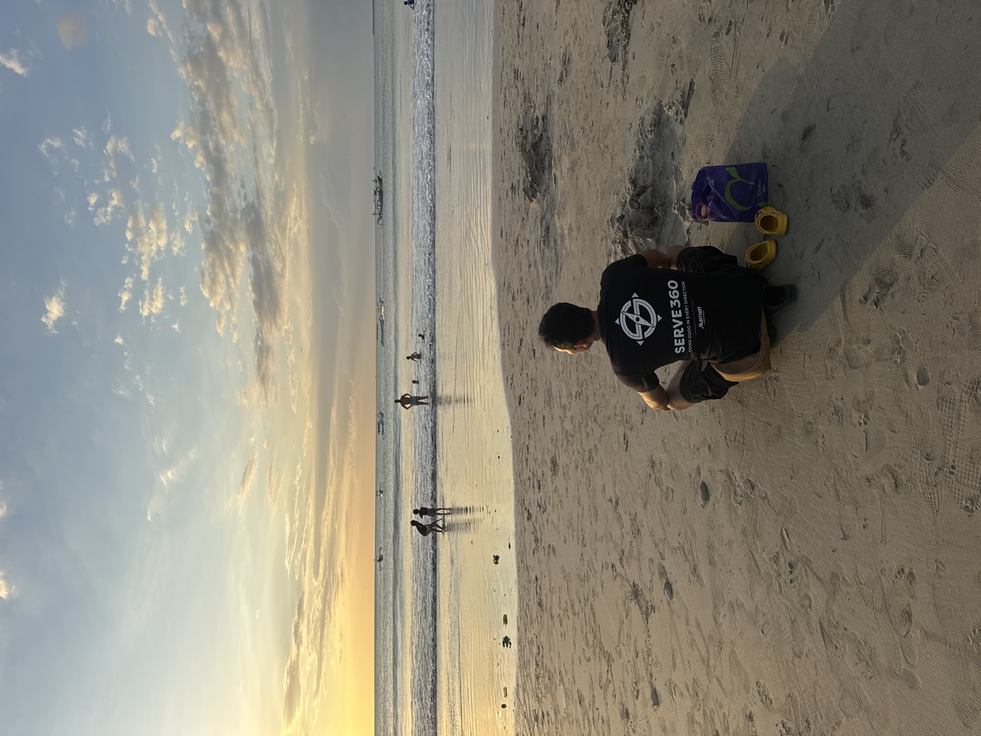 The image depicts a beach in Kuta Selatan, Indonesia at sunset. The sky is predominantly blue with white and golden clouds. The ocean is visible with gentle waves and several people wading in the water. In the foreground, a person wearing a black "Serve 360" t-shirt is kneeling on the sand. Next to them are a purple bag and two yellow shoes. The sand is light in color and covered with footprints. The image is slightly rotated counter-clockwise.