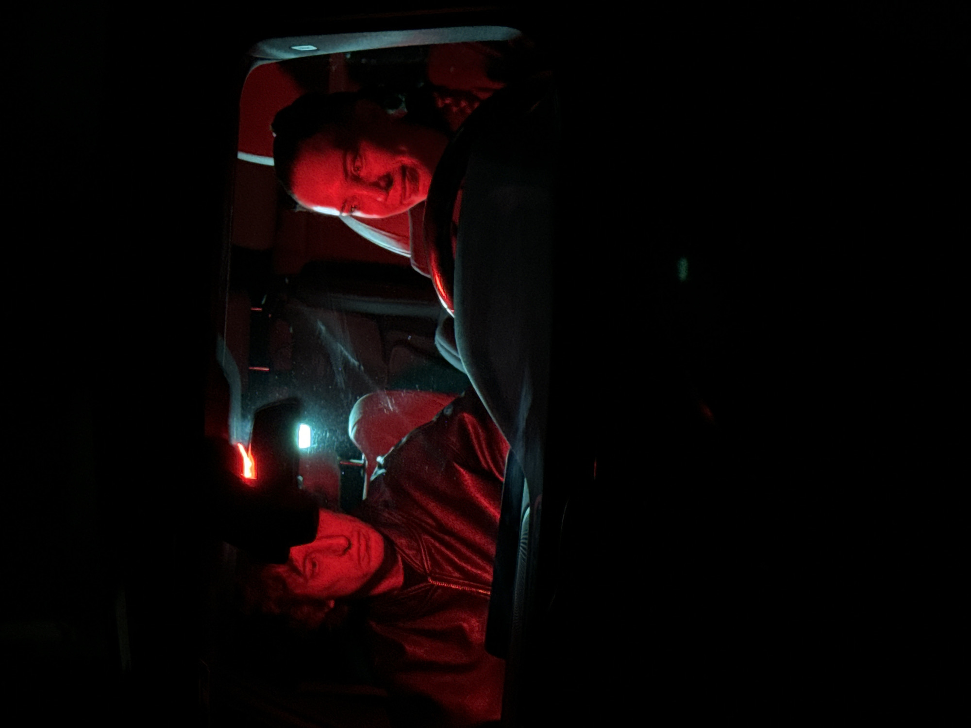 A man lies supine in the back of a car, illuminated by a white light source from the center of the vehicle. His head is visible, facing the rear. A woman, partially visible and closer to the front of the car, looks toward the back. The dominant color is red, likely from an interior light. The environment is dark, the image is focused on the subjects within the car. The location of the scene is not identifiable from the image.