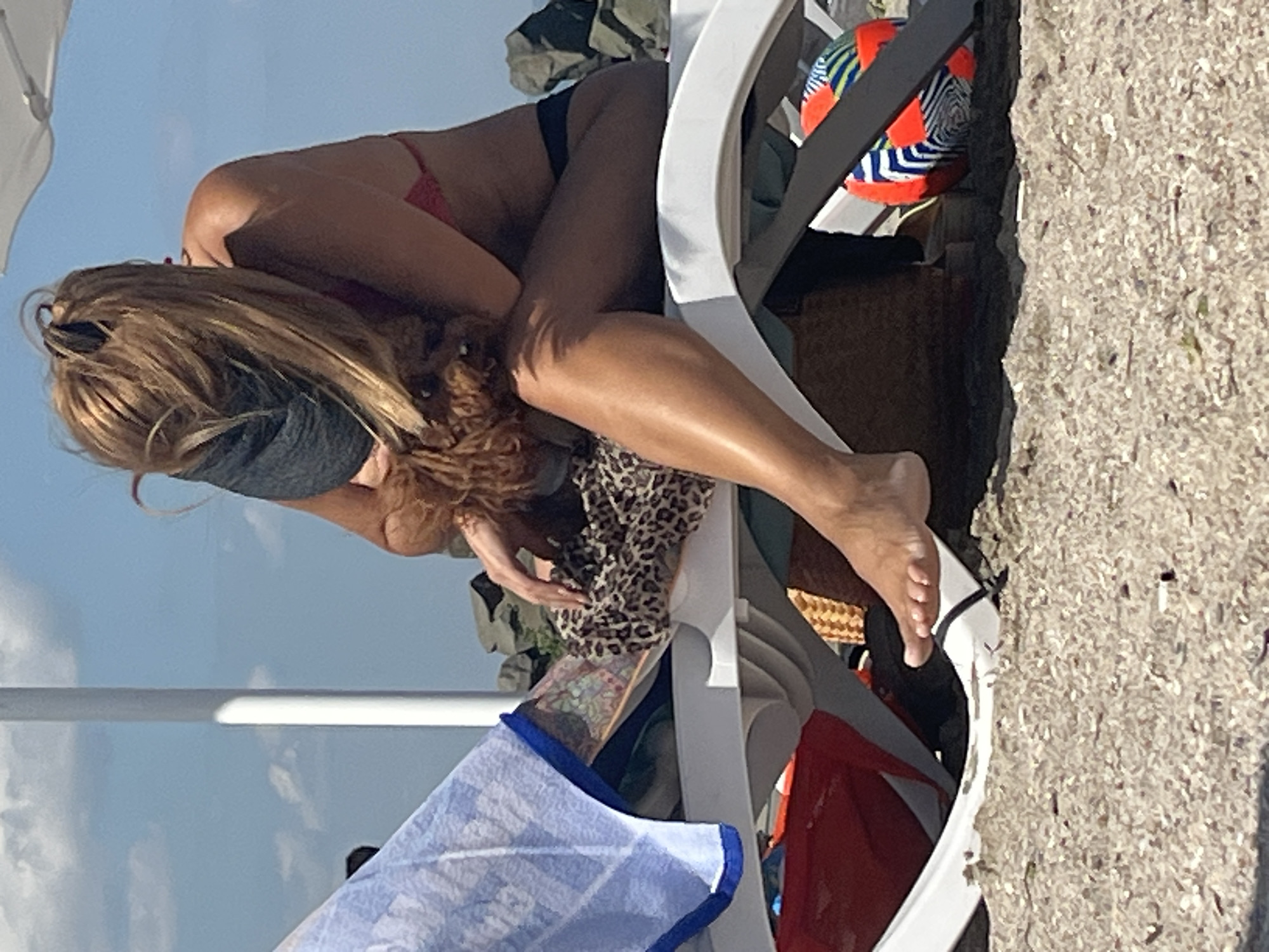 A woman with blonde hair is seated on a white beach chair on a sandy beach. She is wearing a red bikini and a dark visor, and she is holding a small brown dog. To her left, a blue and white patterned towel is draped over the chair. A leopard-print scarf is visible near the dog. In the background, there are large rocks and a clear blue sky with some clouds. A colorful ball with geometric patterns is on the sand to the right of the chair. The scene appears to be on a beach in Pomorie, Bulgaria.