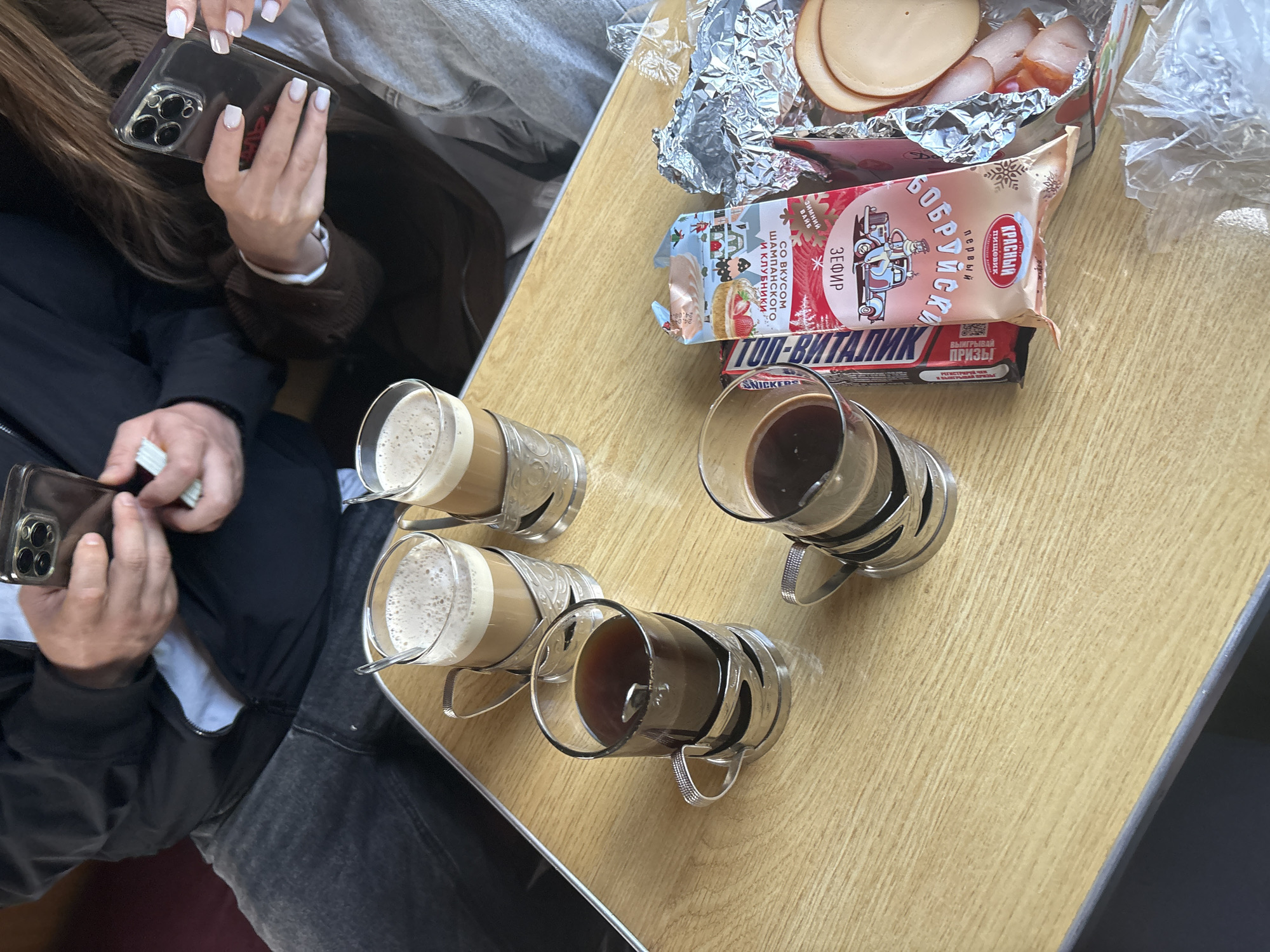 The image shows a table with four beverages in glass mugs and food items. Two of the beverages appear to be coffee with milk and the other two appear to be coffee. Food includes a foiled tray of sliced meat, a package labeled "Бобруйска" (Babruysk), and a Snickers bar. Two people are present, both holding smartphones; one is holding what appears to be money. This scene may be located in a train or other travel conveyance.
