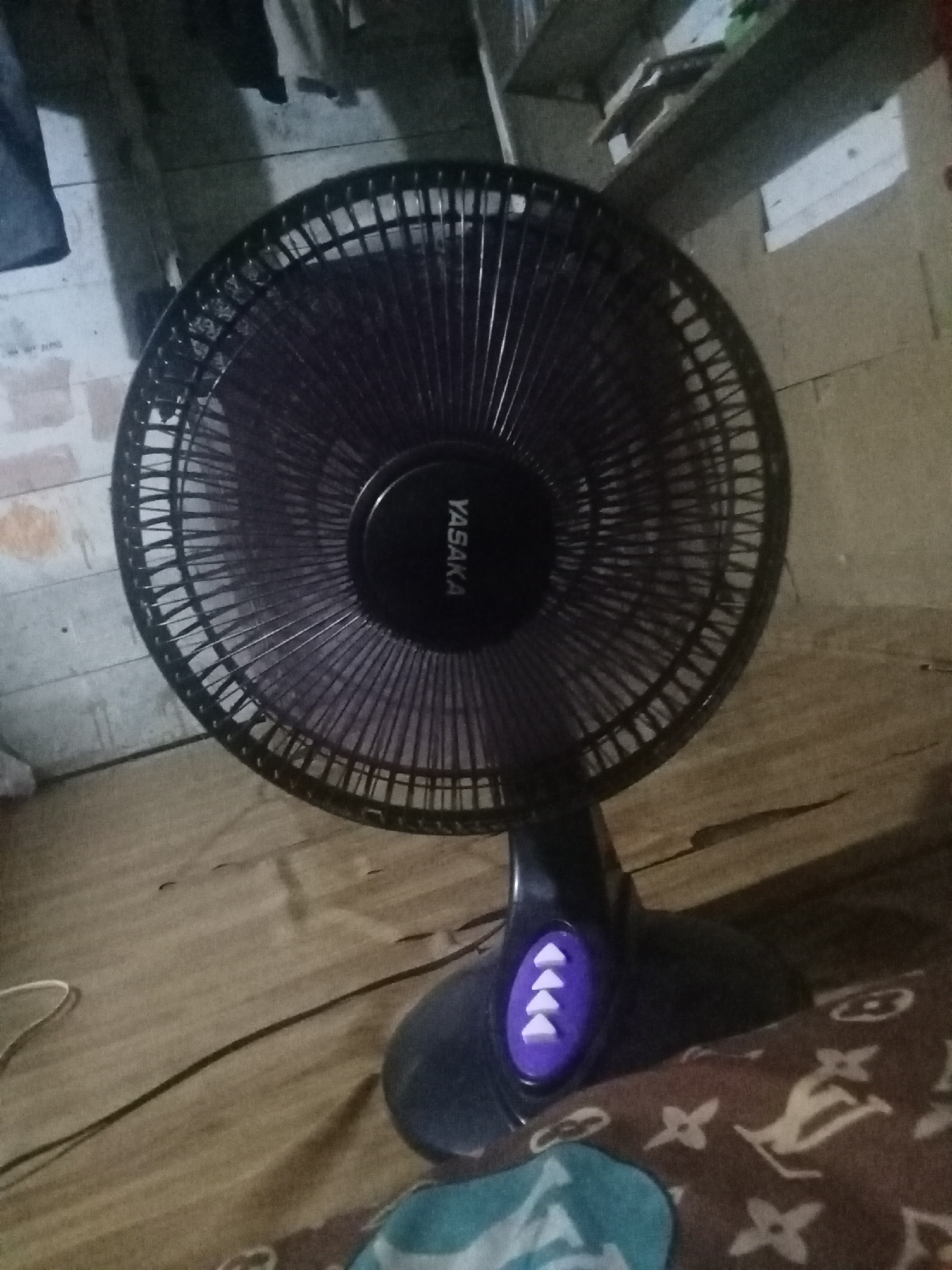 A black oscillating fan with a "YASAKA" logo on its motor housing is positioned on a wooden floor covered with a patterned blanket. The fan has a dark grille and a black base with purple buttons. In the background, there is a wall with a shelf and some hanging items. The scene appears to be indoors, likely in a residential setting in Buketglumpang, Indonesia.