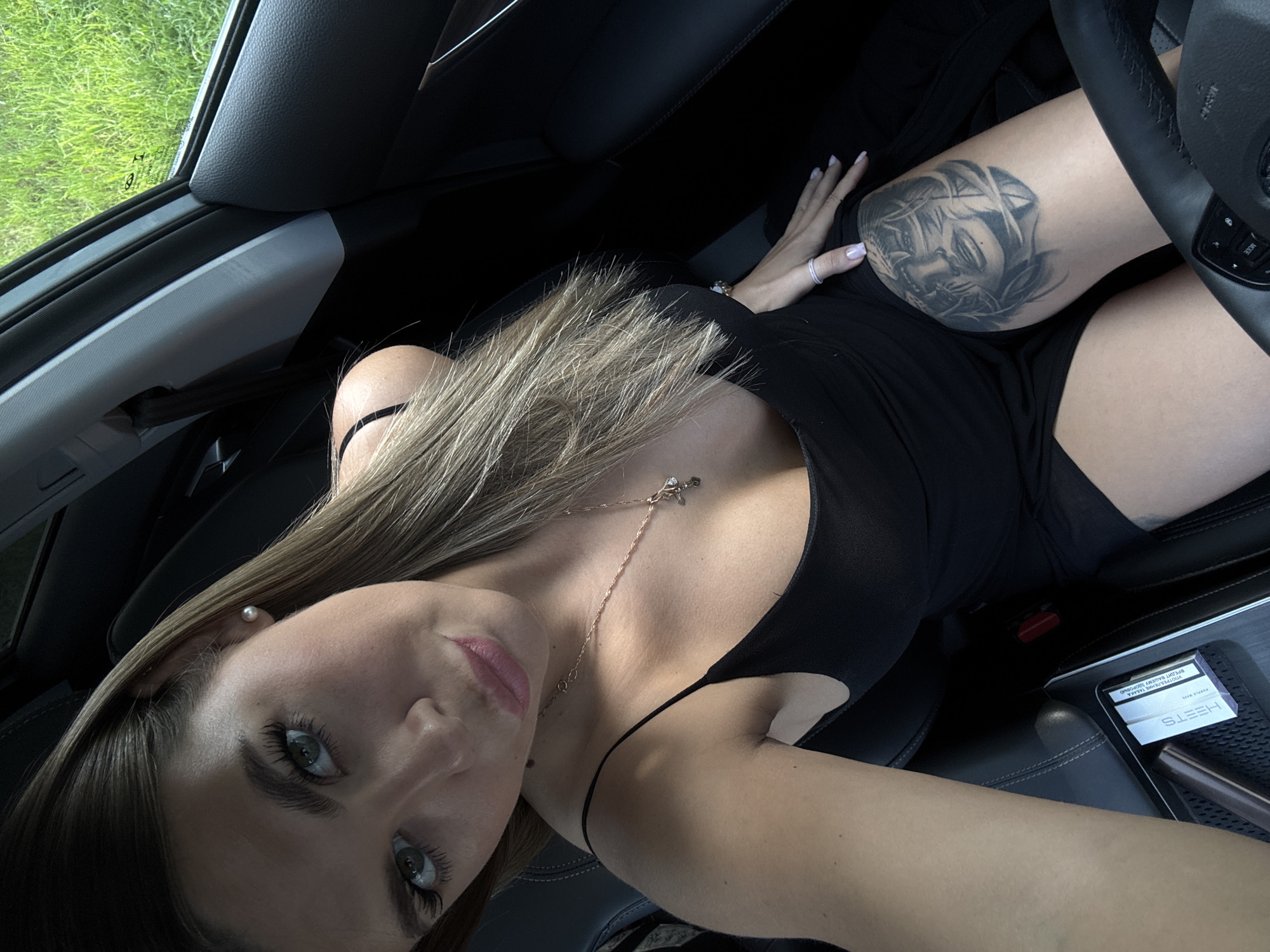 A woman with blonde hair and green eyes is sitting in the driver's seat of a car. She is wearing a black spaghetti strap dress and a necklace with a pendant. She has a tattoo of a woman's face on her thigh. The car is parked outdoors, with a grassy field visible through the window. A pack of HEETS tobacco sticks is on the center console.